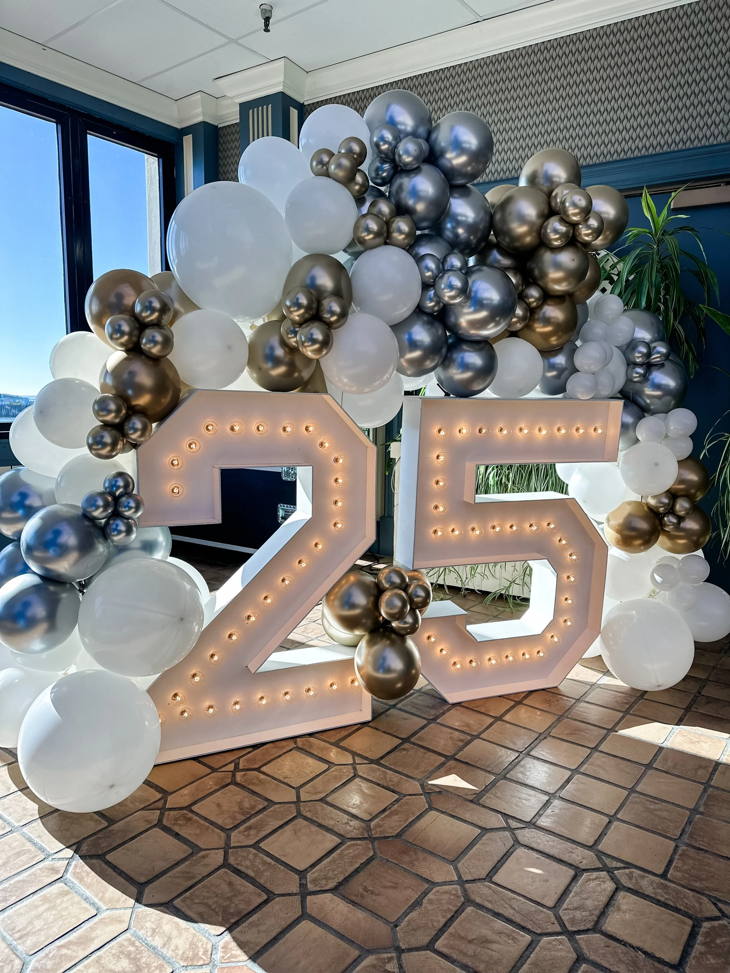 Decorative 25th celebration display featuring large illuminated numbers '25' surrounded by white, gold, and silver balloons arranged in an arch.