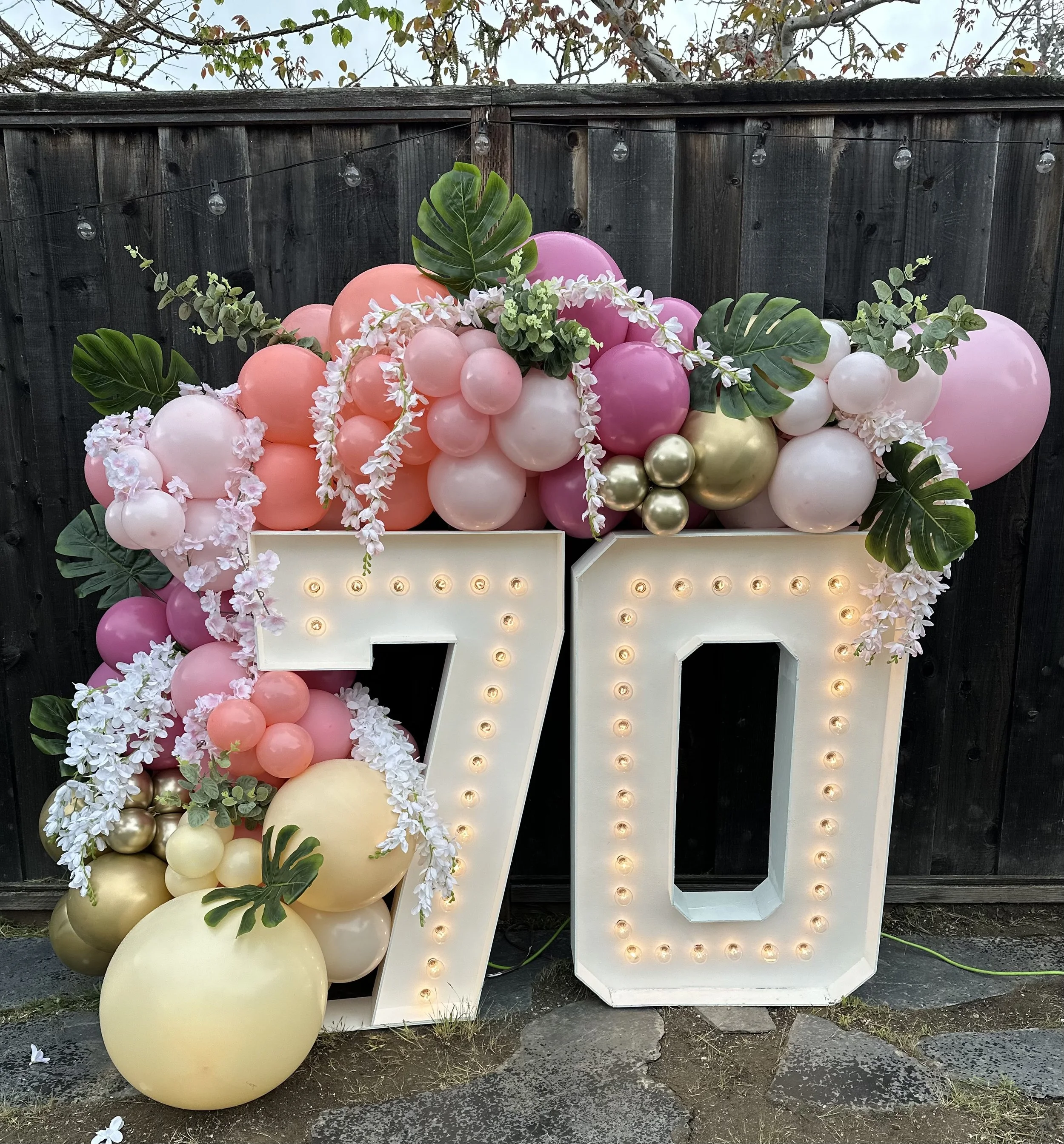 Large illuminated numbers '70' decorated with pink, white, and gold balloons, green leaves, and white flowers, set against a dark wooden fence with string lights above.