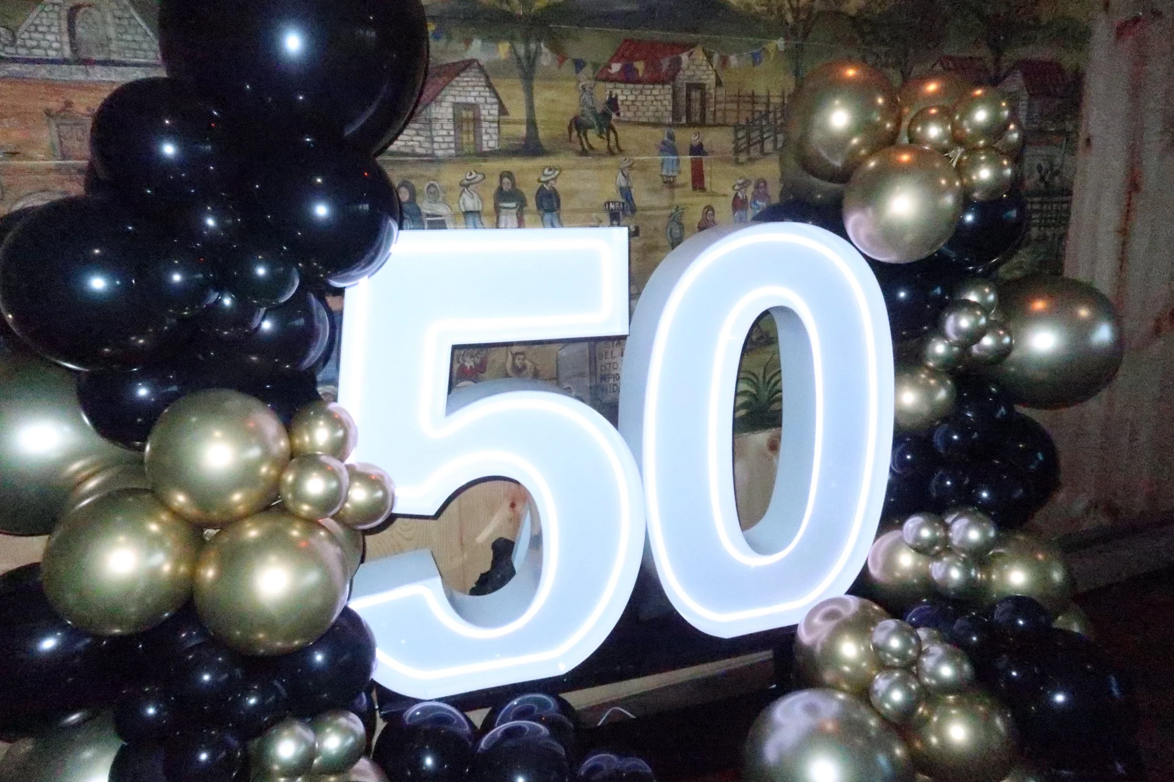 Large illuminated number 50 surrounded by black and gold balloons, set against a colorful mural background.
