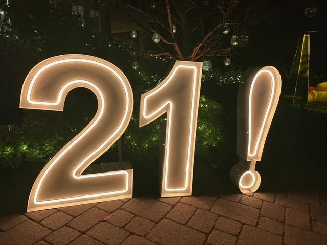 Large illuminated signs spell out '21!' on a brick paved ground at night.