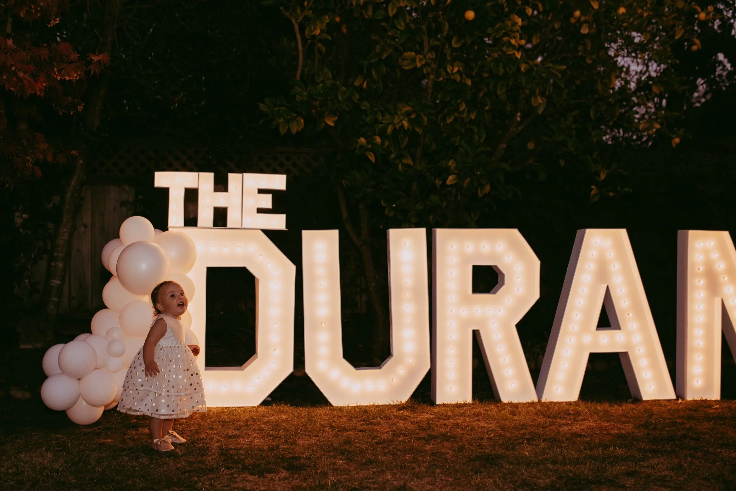 A young girl in a white dress standing next to large illuminated letters spelling 'DURAN' and decorative white balloons at night time.
