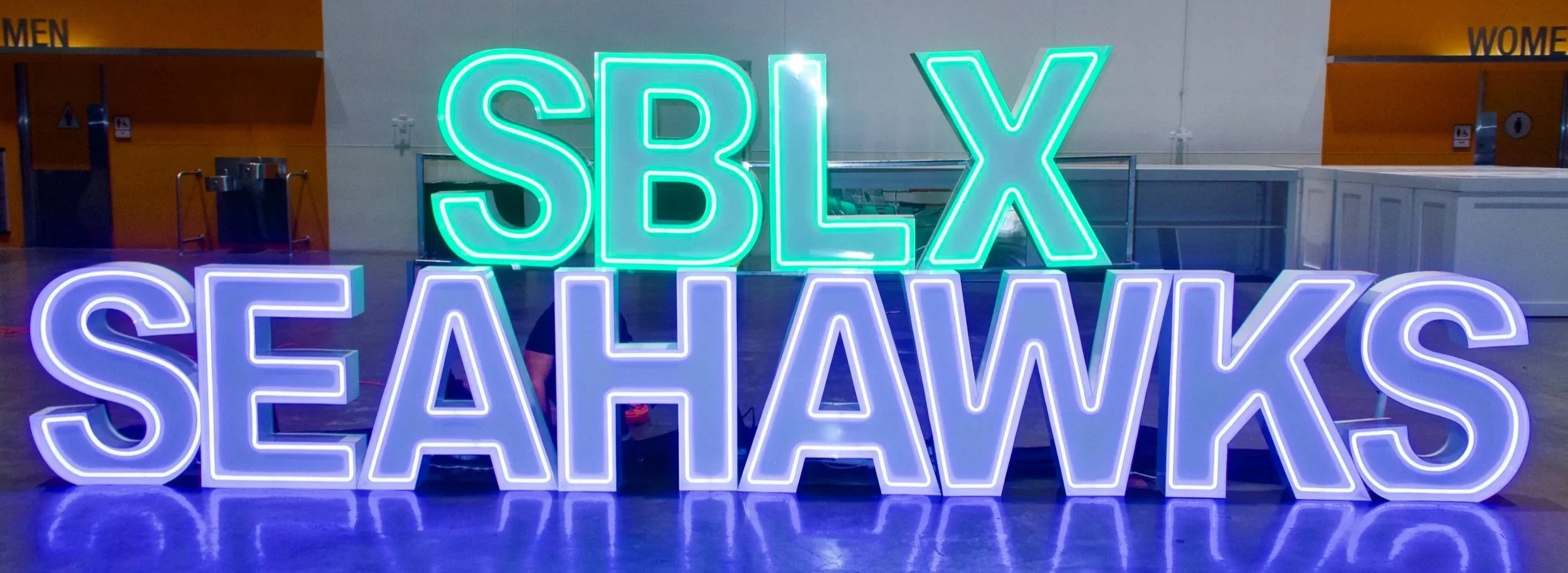 Neon sign that reads 'SBX SEAHAWKS' with 'SBX' in green and 'SEAHAWKS' in purple, displayed indoors with a background of orange and white walls and doors indicating men's and women's restrooms.
