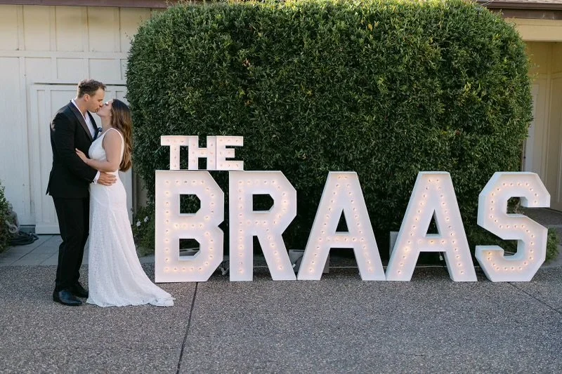 A couple in wedding attire standing close and embracing each other in front of large illuminated marquee letters spelling 'THE BRAAAS' on a paved outdoor surface with a bush in the background.