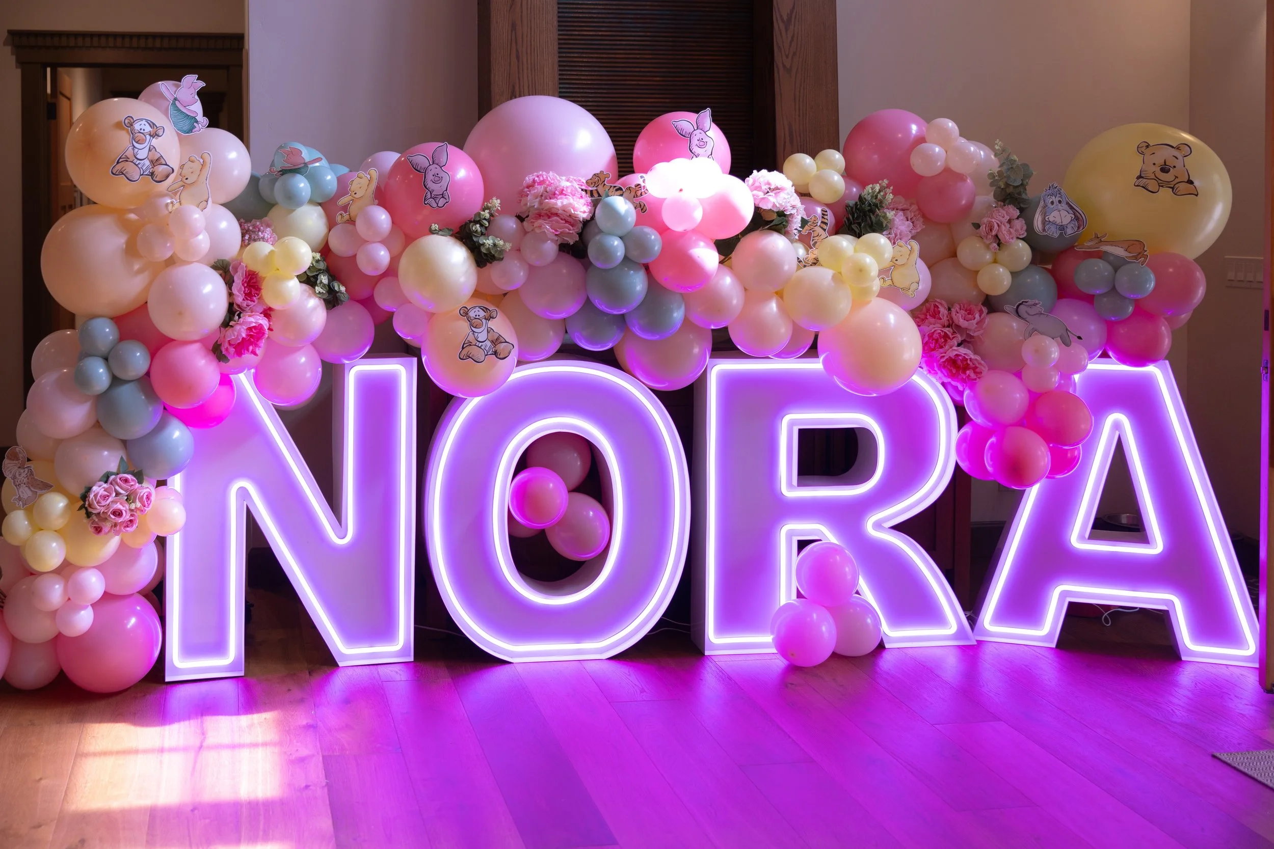 Neon lit sign spelling 'NORA' surrounded by pink, yellow, and blue balloons with cartoon animal and character stickers, along with pink flowers, set against a wooden wall and floor.