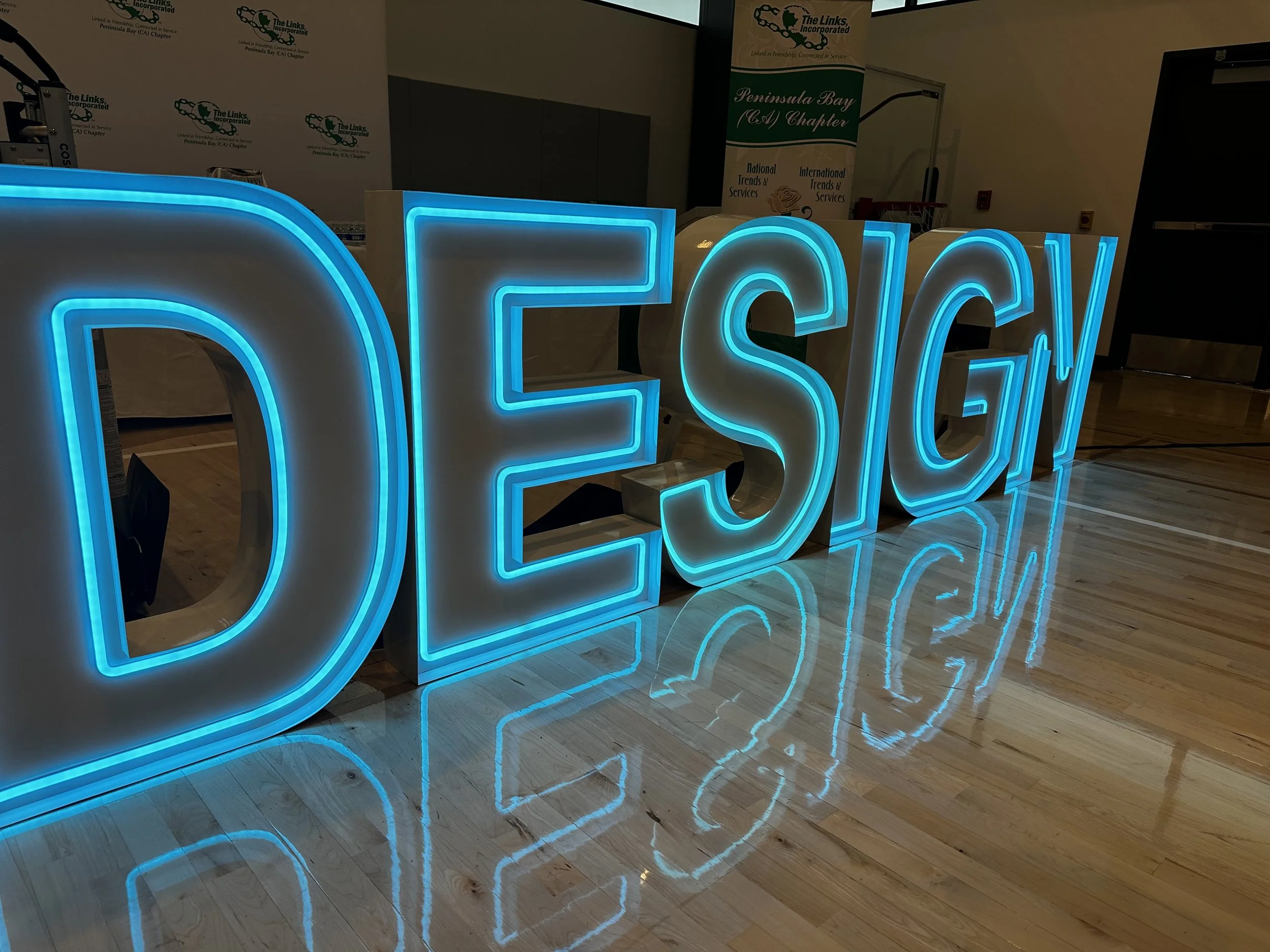 Large illuminated sign spelling out "DESIGN" with blue neon outline, reflected on a polished wooden floor. Event banners in the background mention "Peninsula Bay (A) Chapter" and "The Links, Incorporated."