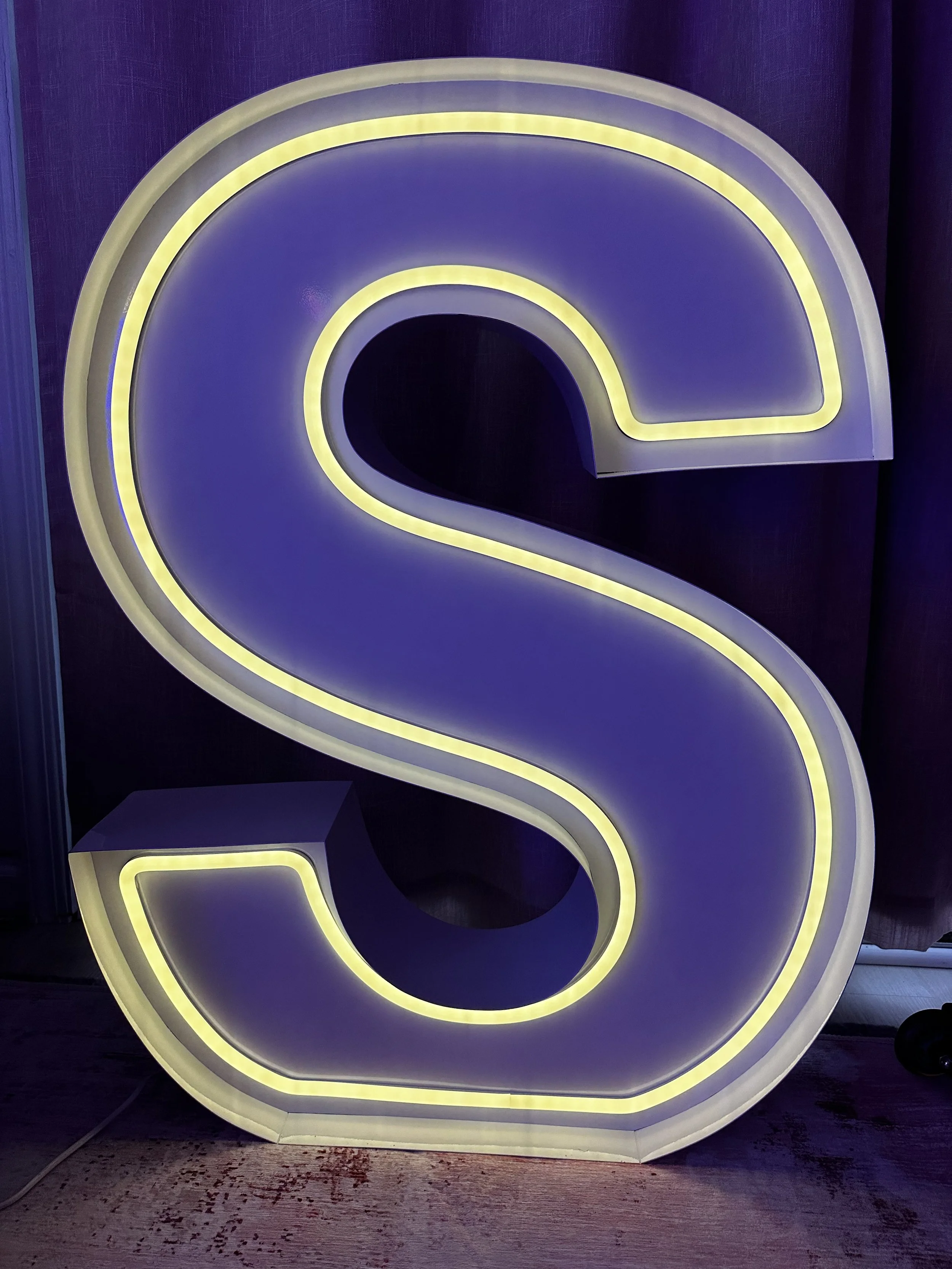 Illuminated purple and yellow neon letter 'S' sign against a dark background.