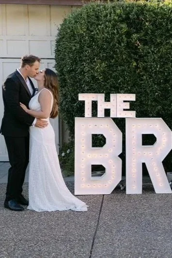 A couple in wedding attire kissing outdoors next to large illuminated letters spelling "THE BR" with a green bush in the background.