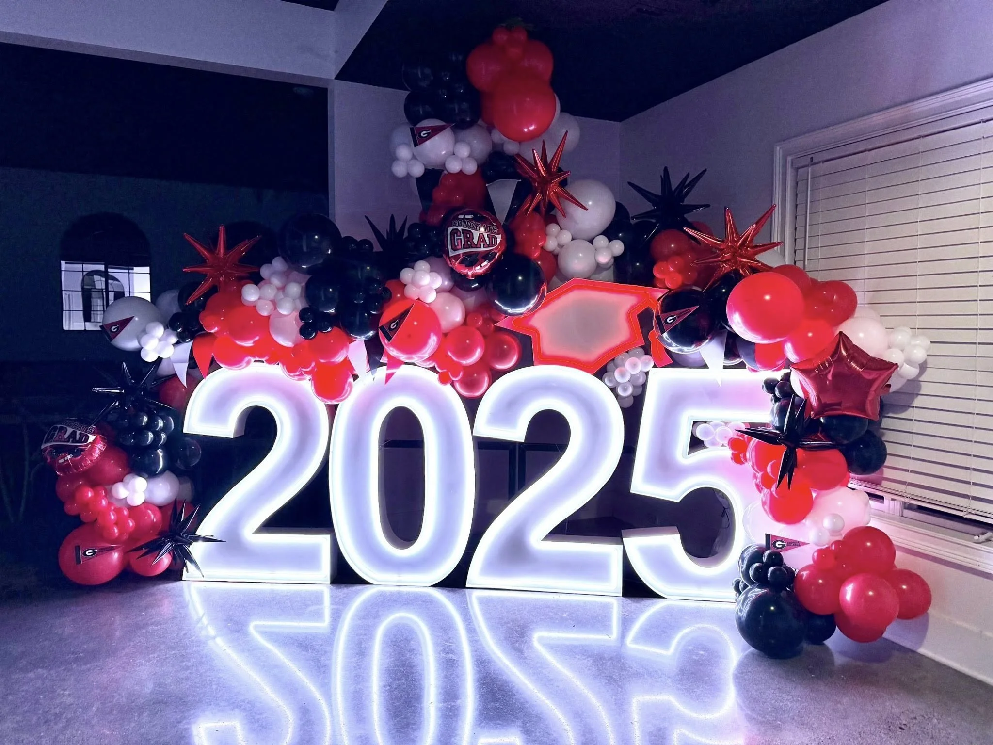 Decorative 2025 graduation celebration balloons in black, red, and white, including large illuminated number signs '2025' and various shaped balloons, some featuring the Georgia Bulldogs logo, arranged in a festive display inside a room.