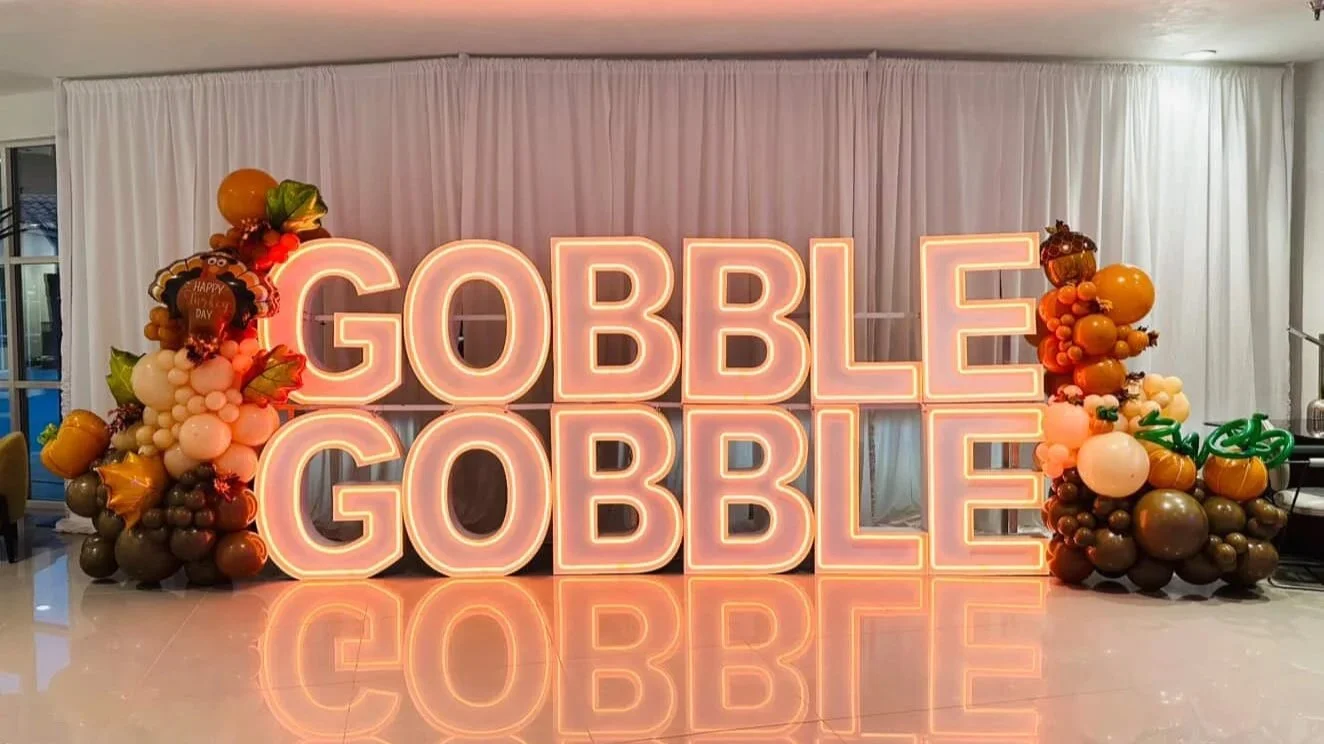 Decorative fall-themed balloon arrangement surrounding a large illuminated sign that reads 'GOBBLE GOBBLE' with white curtains and a reflective floor