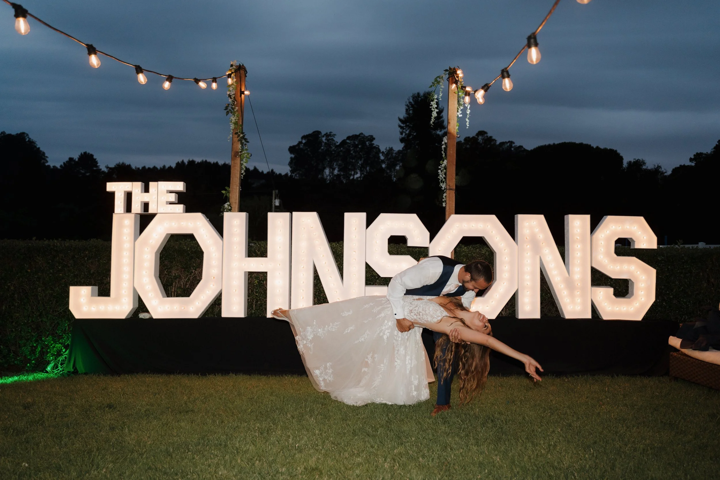 A couple dancing at their wedding reception outdoors with illuminated sign reading 'The Johsons' in the background, string lights overhead, and darkening evening sky.
