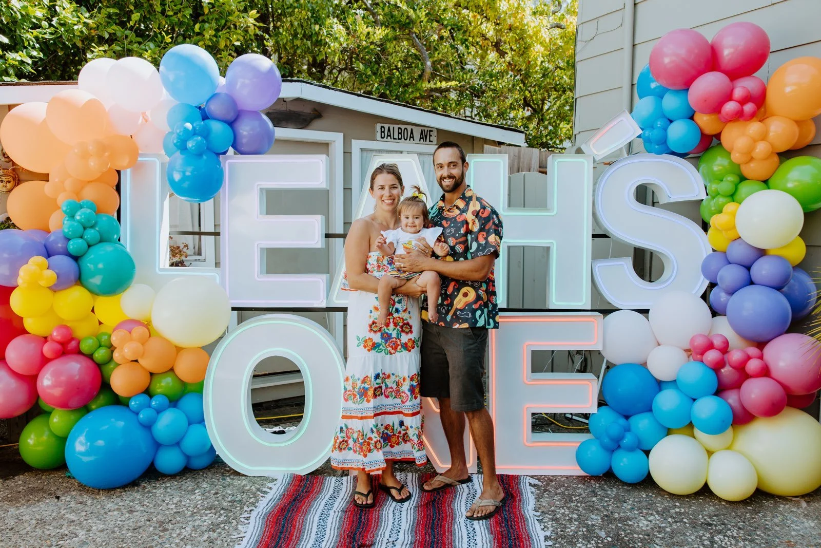 Family of three standing in front of a colorful balloon display spelling 'LOVE' at an outdoor celebration, with a woman holding a young girl and a man standing beside them.