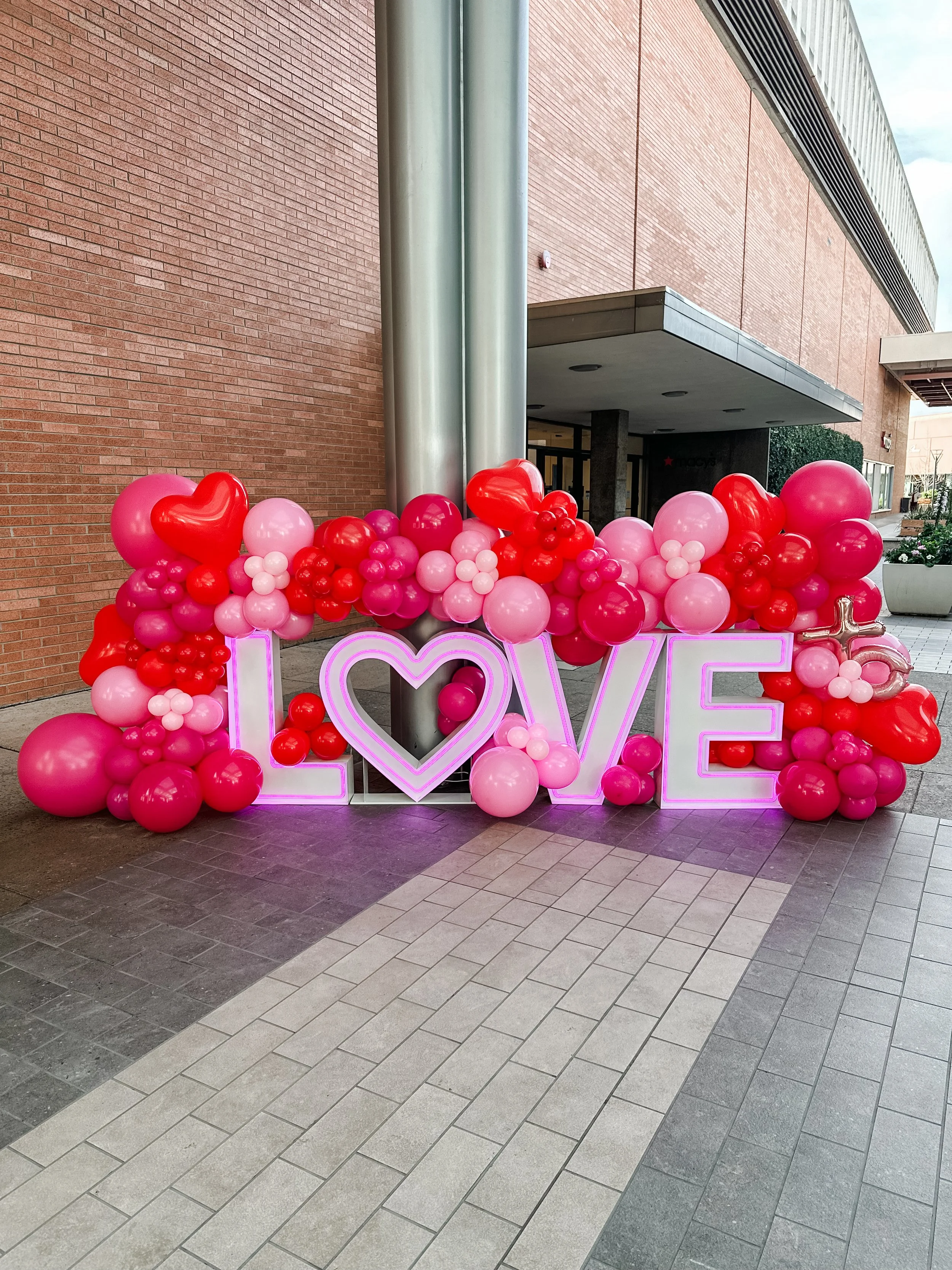 Decorative installation with pink and red balloons and a neon sign spelling 'LOVE' outside a building.
