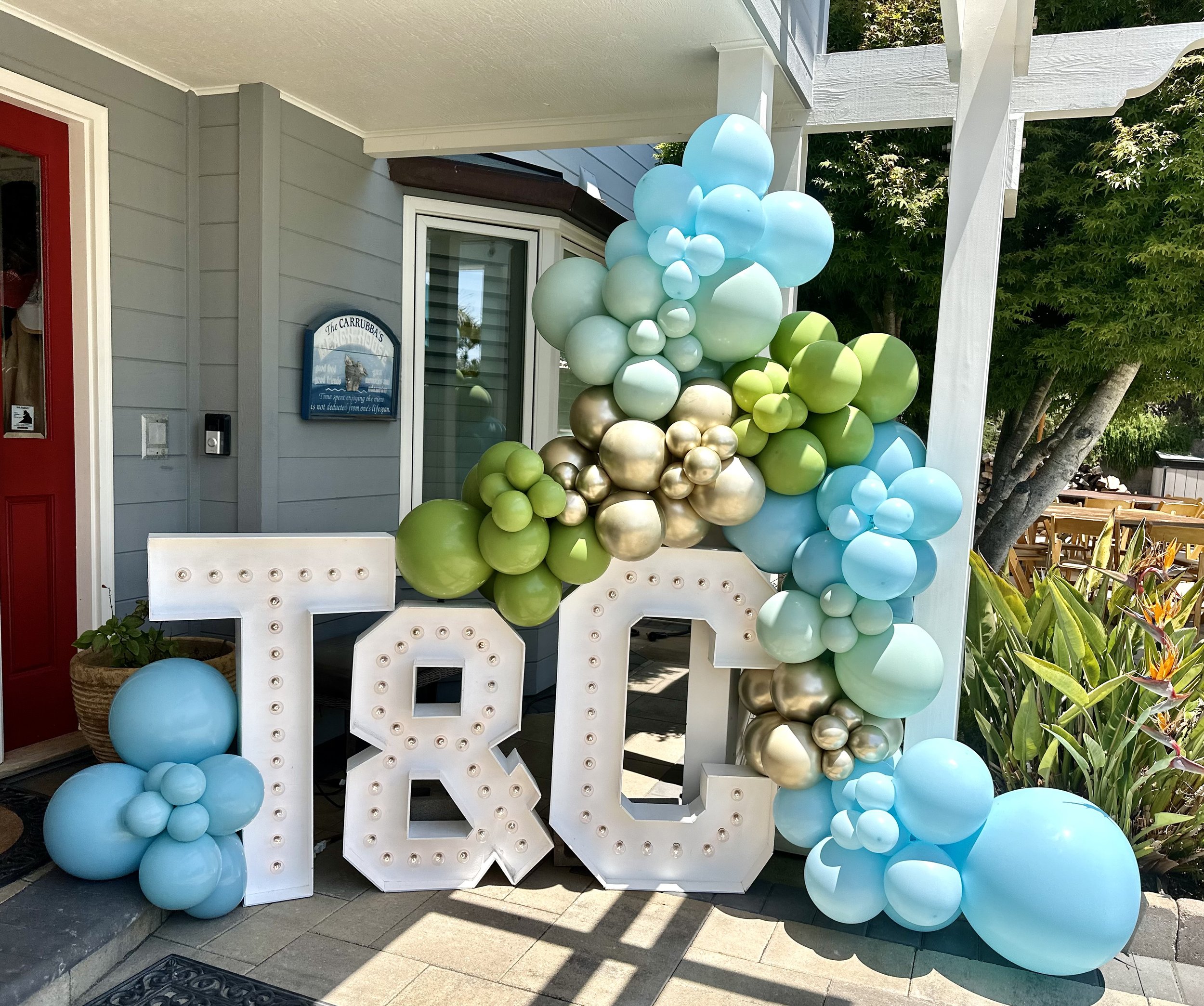 A decorative display on a porch with large white letters spelling 'TOQ' and a cluster of pastel-colored balloons in shades of blue, green, gold, and silver surrounding the letters.