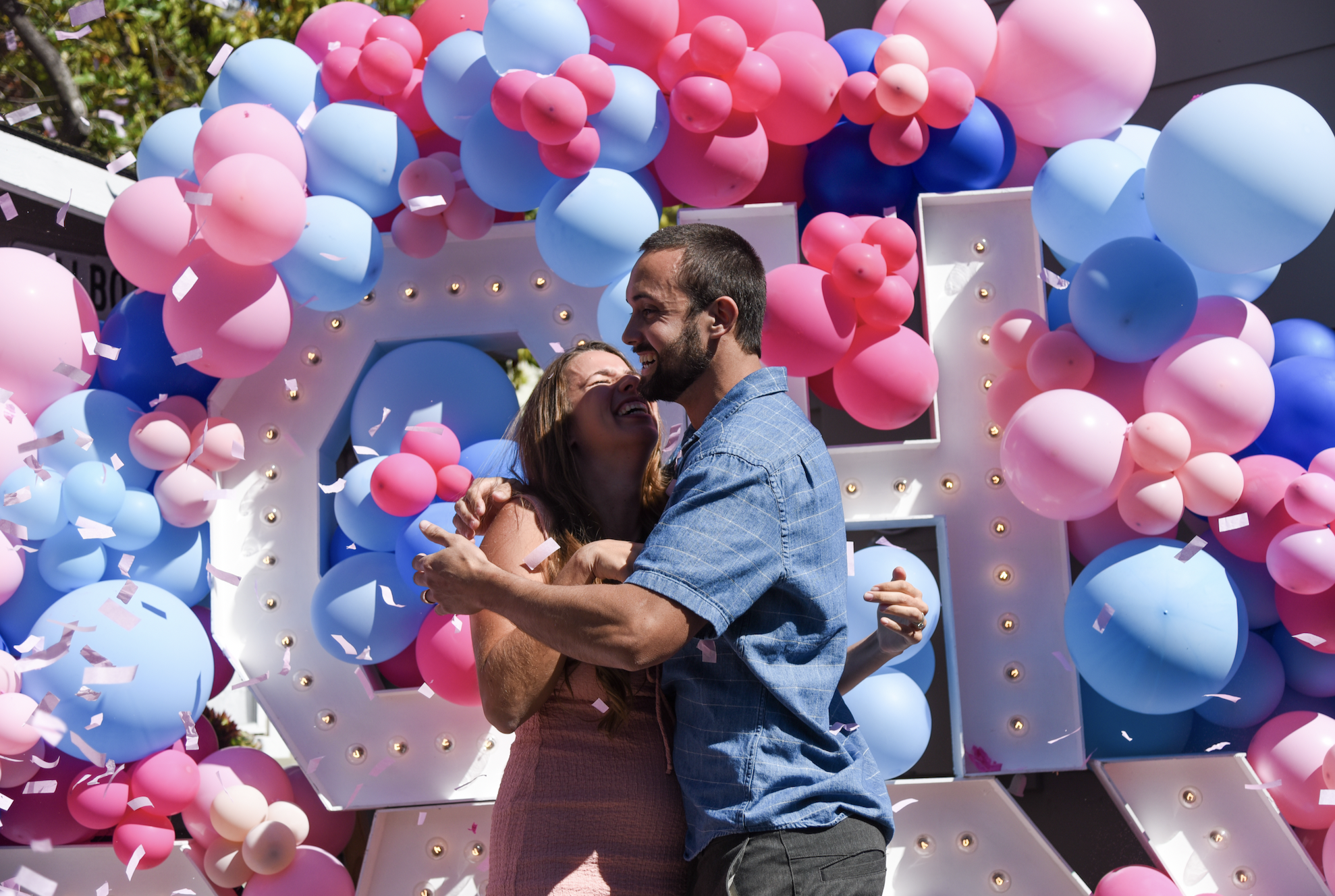 A couple is hugging and smiling in front of a large decorative backdrop made of pink, blue, and purple balloons and a white marquee letter "O" with small light bulbs.