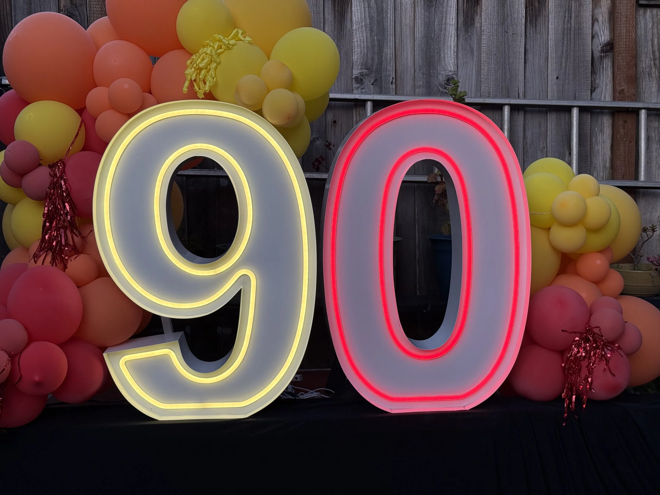 Neon signs displaying the numbers 90 in yellow and red, surrounded by balloons in shades of pink, orange, and yellow, with a wooden fence in the background.