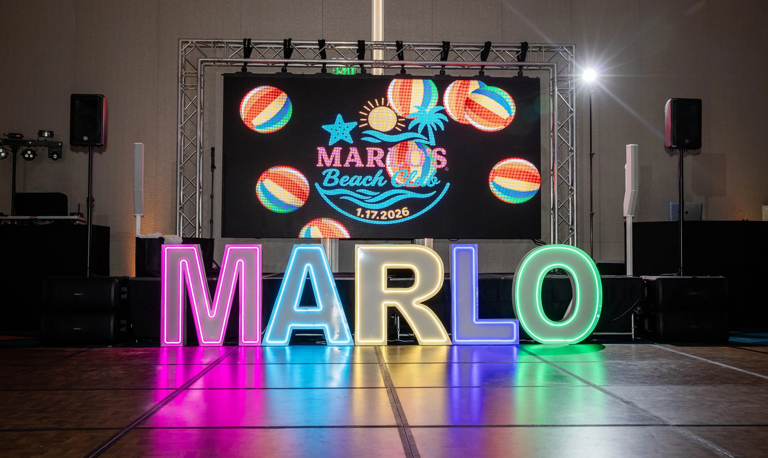 Colorful neon letters spell out 'MARLO' on a stage with a large screen behind displaying a beach-themed event, 'Marlo's Beach Club,' with graphics of balls, sun, starfish, and date January 17, 2026.