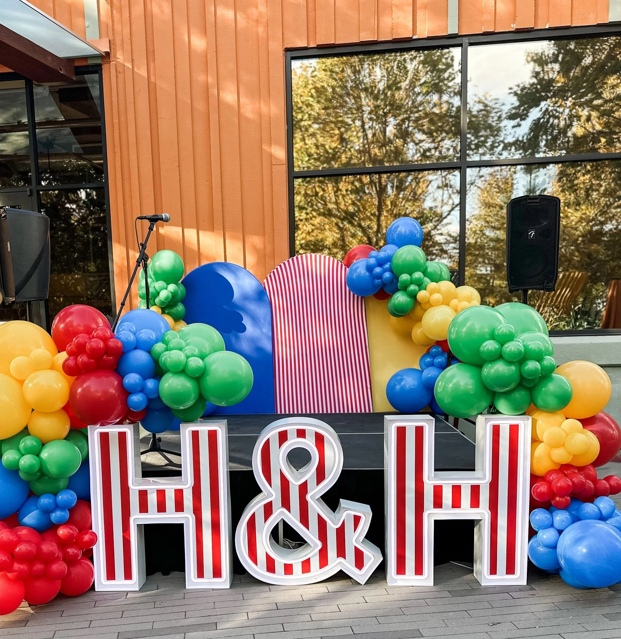 Decoration for a celebration with large letters 'H&L' in red-and-white stripes, surrounded by colorful balloons in green, yellow, red, and blue, and striped fabric backdrop behind a small stage with microphone and speakers.