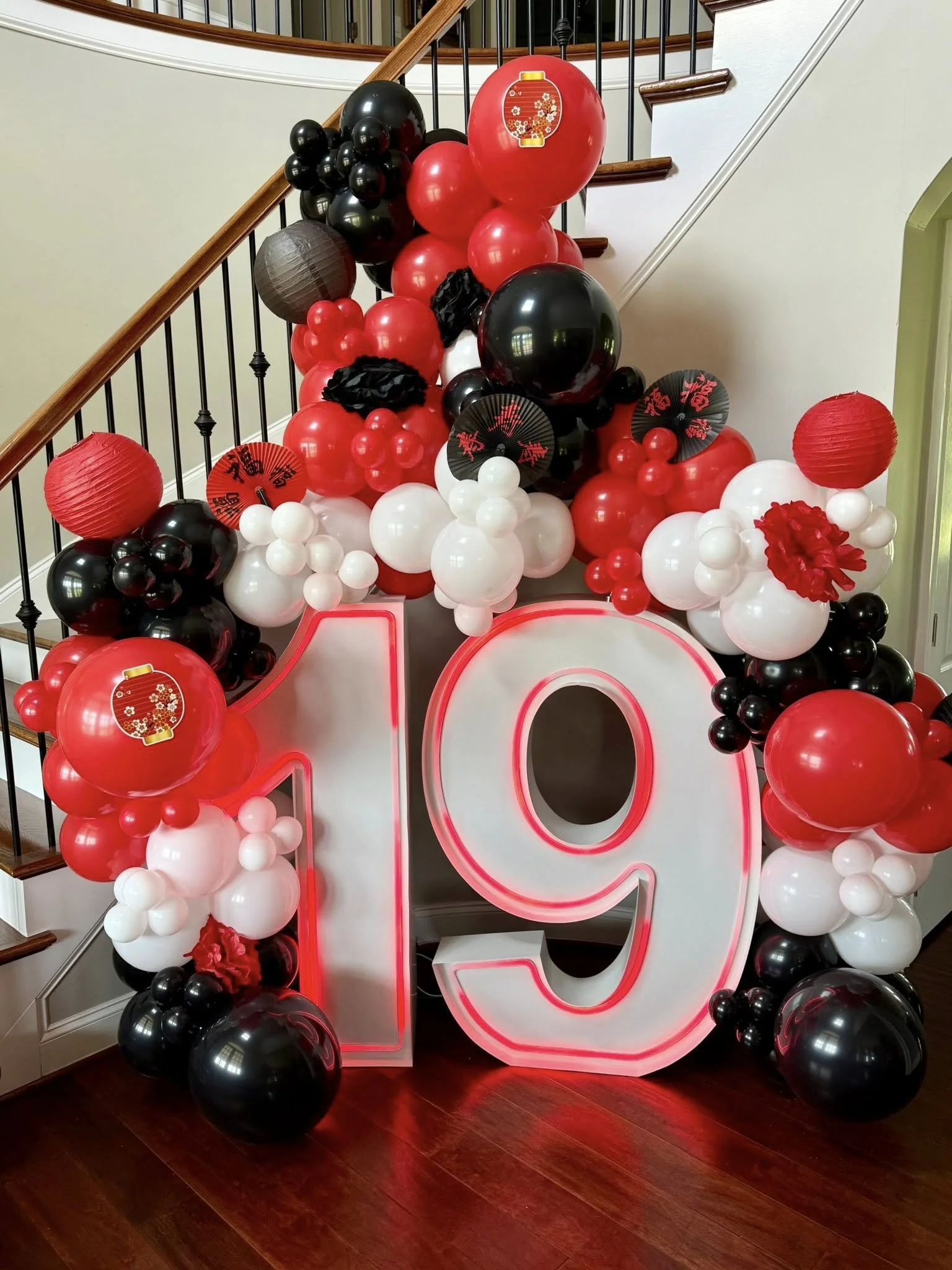 Decorative birthday display with a large illuminated number 19, surrounded by red, black, and white balloons, some with paper fans, on a staircase landing.