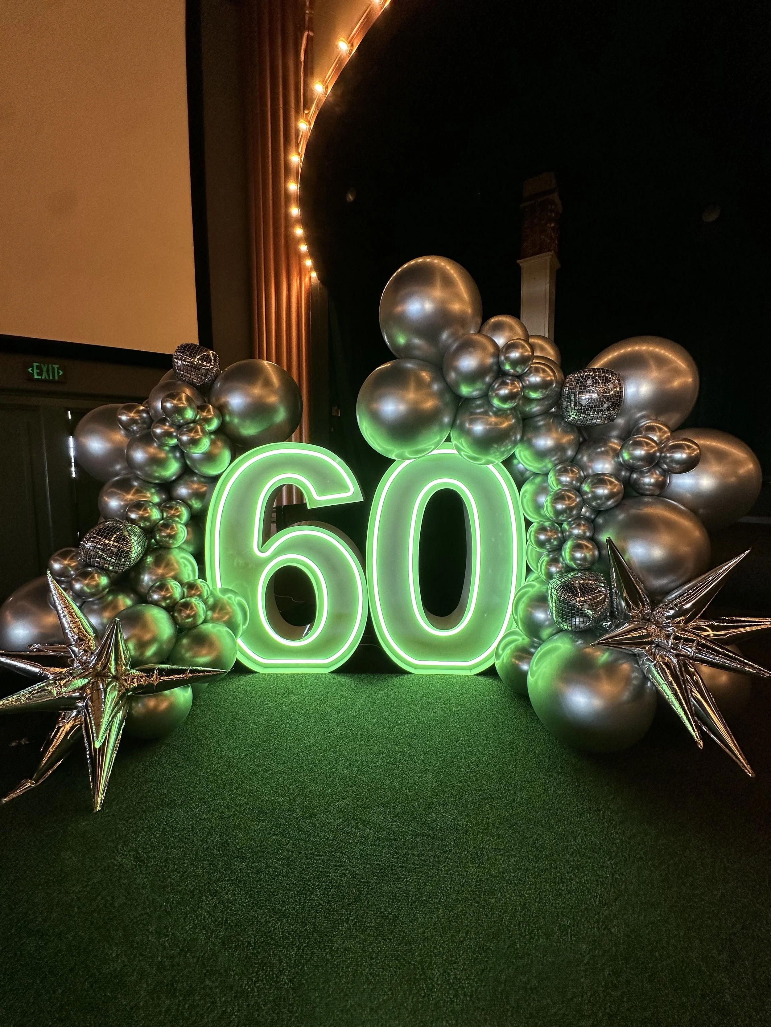 Decorations celebrating a 60th birthday with a green illuminated '60' sign surrounded by balloons and metallic star-shaped balloons on a green carpet.