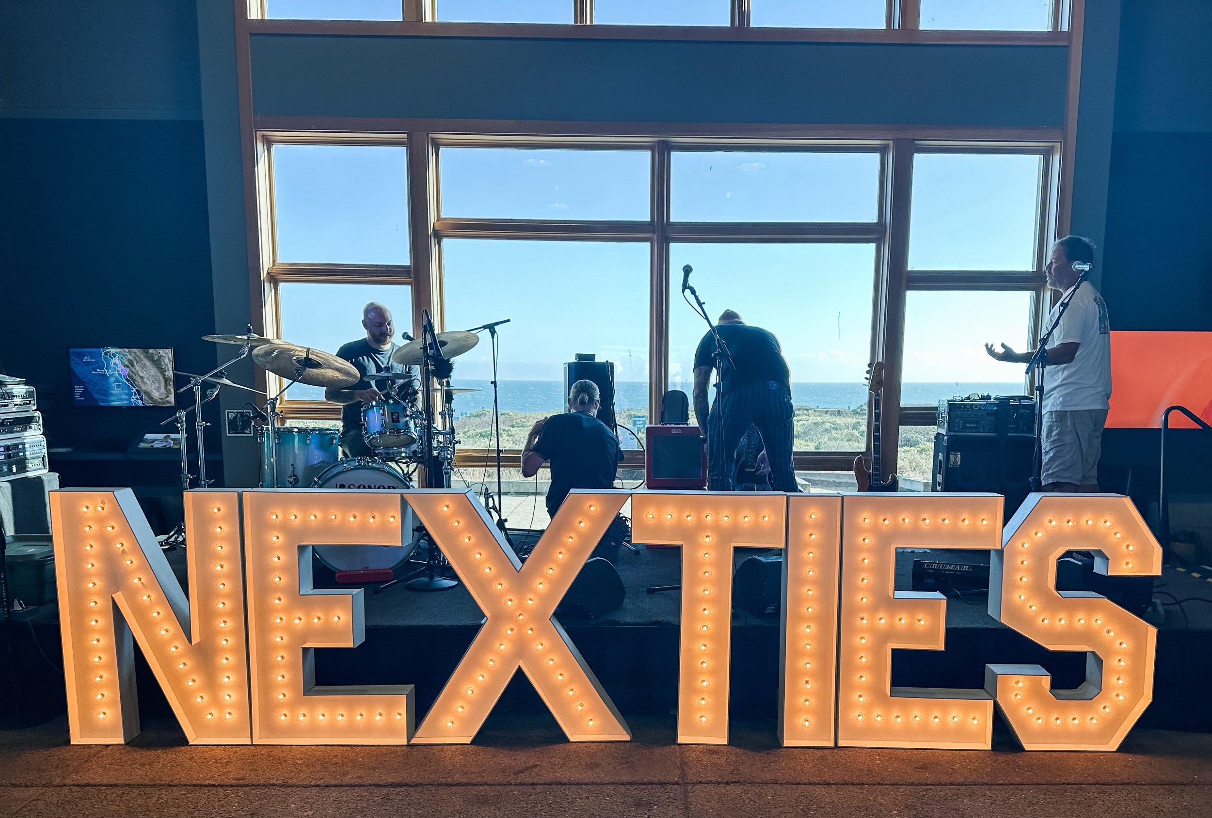 A band setup performs on a stage with large illuminated sign spelling 'NEXITIES' in front. The stage is indoors with large windows showing an ocean view. The band includes at least three members: a drummer, a person with a microphone, and a person wi