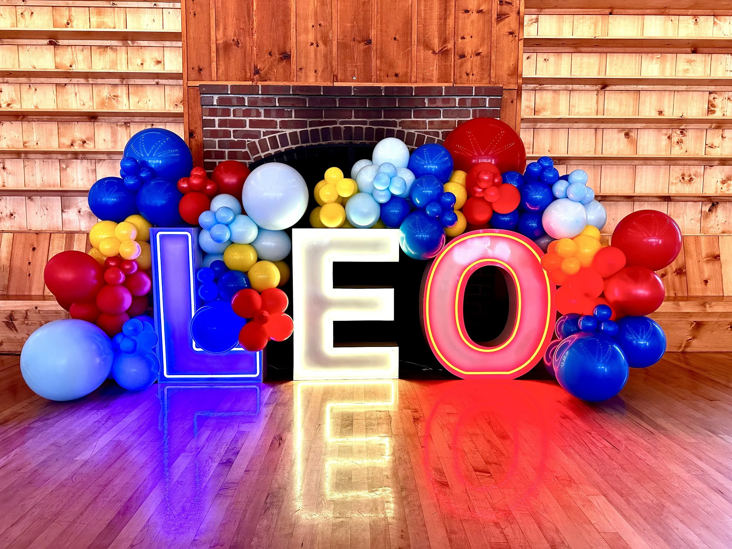 Colorful balloons arranged around large illuminated letters spelling 'LOVE' in front of a brick fireplace with a wooden wall background.