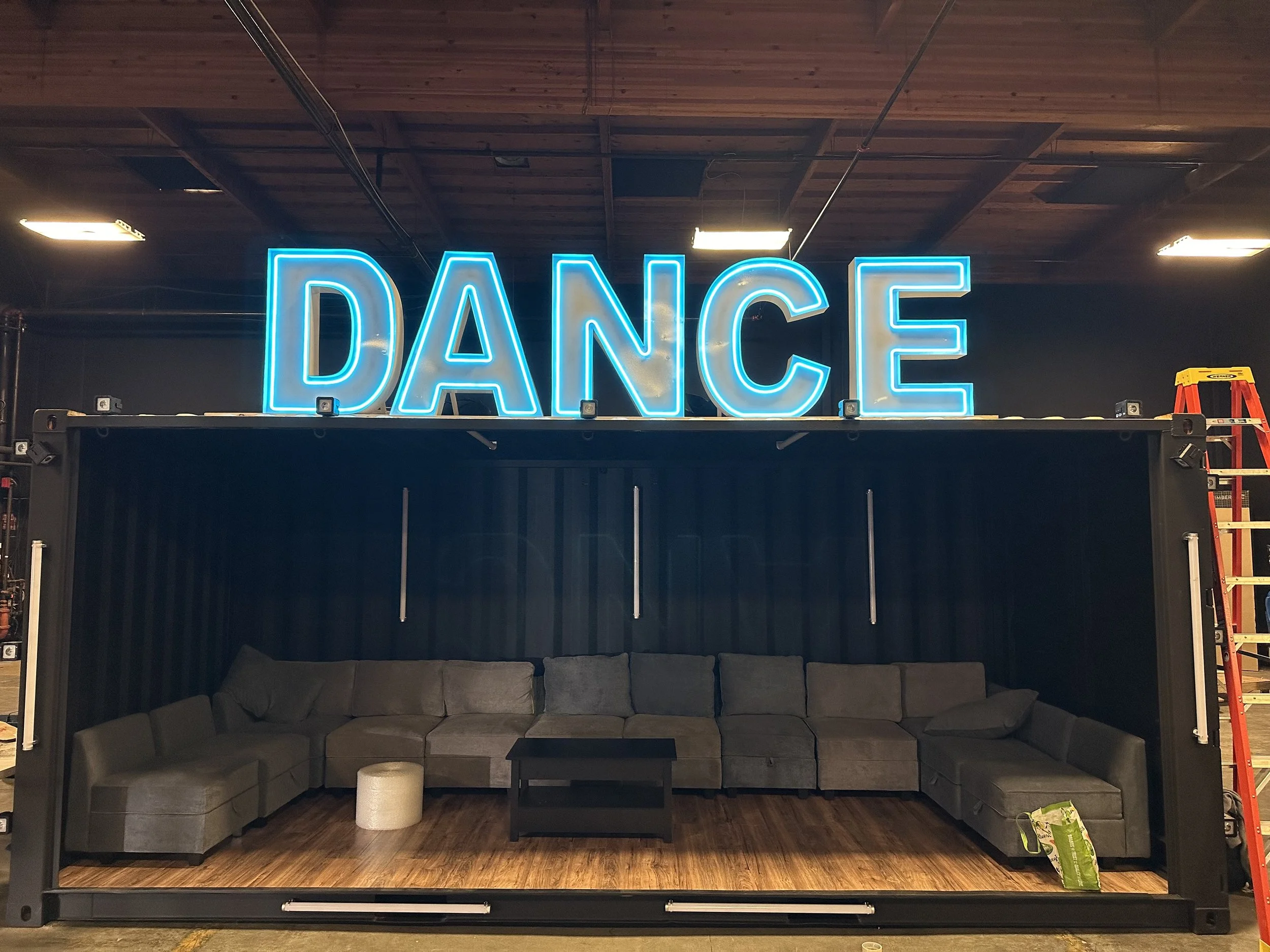 A neon sign spelling 'DANCE' mounted above a black, shipping container-style room with a curved gray sectional sofa and a small black table inside, set on a wooden floor.