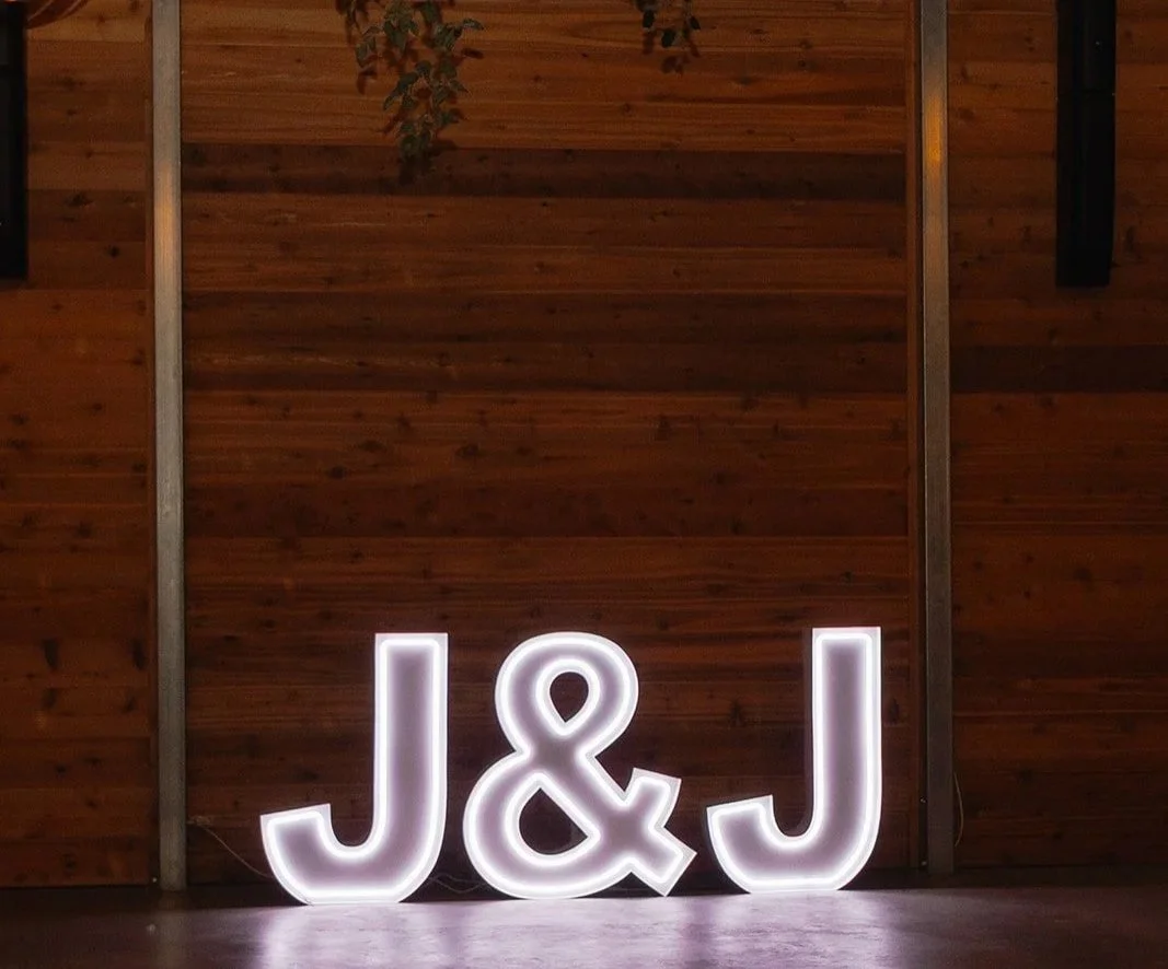 Illuminated white neon letters displaying "J&J" on a wooden wall.