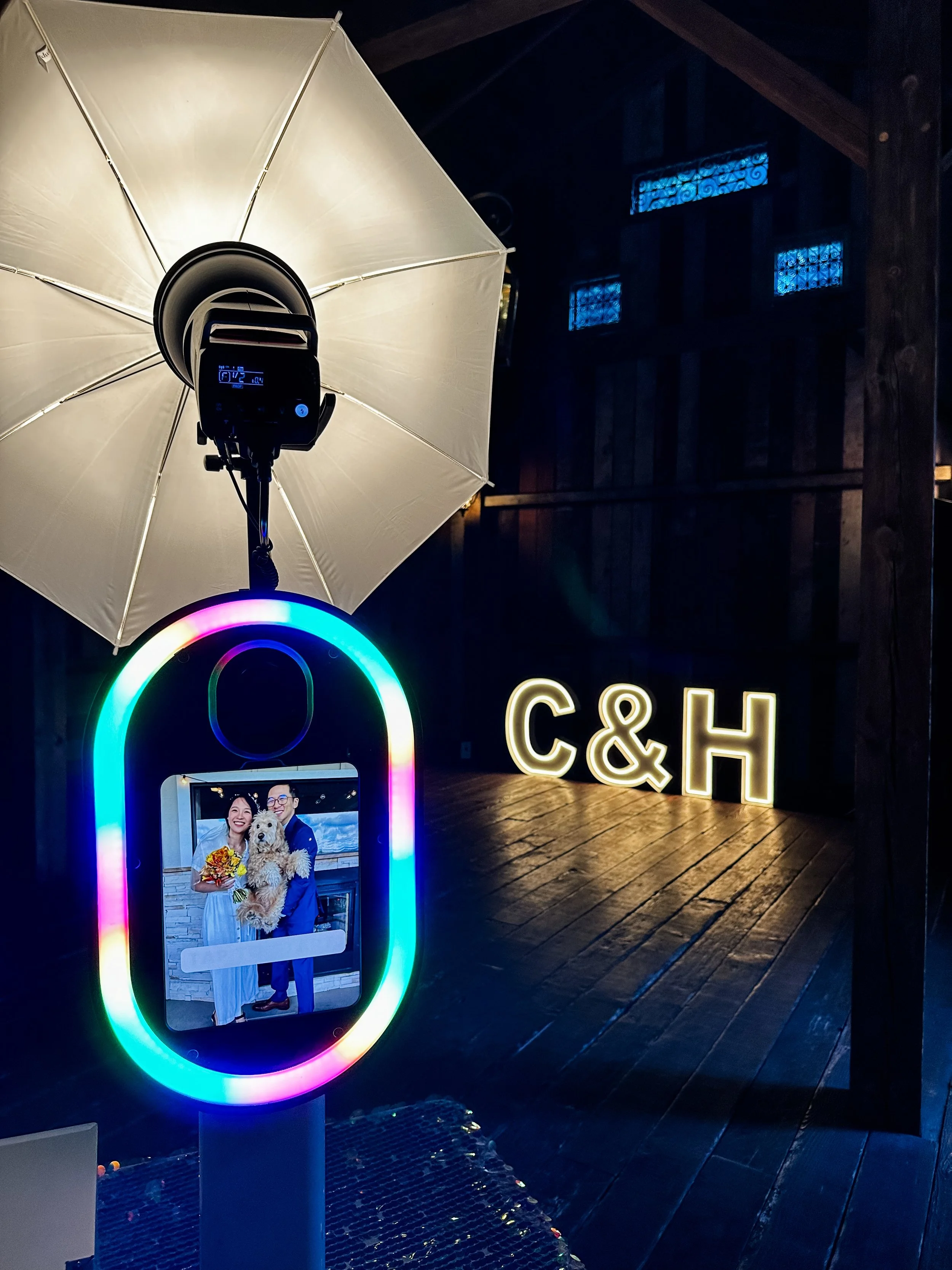Photo of a digital photo booth with a smiling couple holding a dog, illuminated by colorful LED lights, with a neon sign reading 'C&H' in the background.