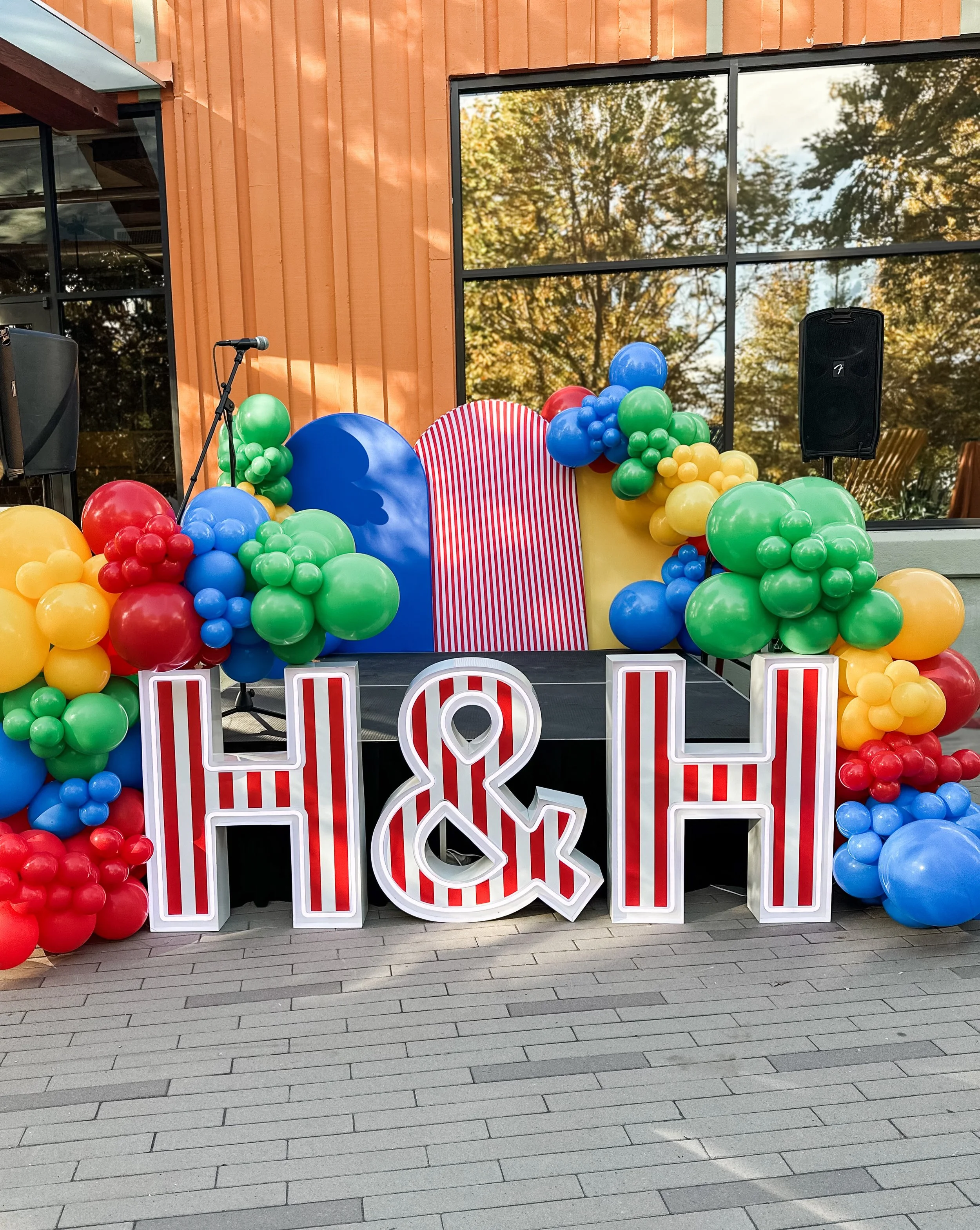 Colorful balloon decorations with large illuminated letters spell out 'H& H' at an outdoor event with a wooden building and trees reflected in the glass window.
