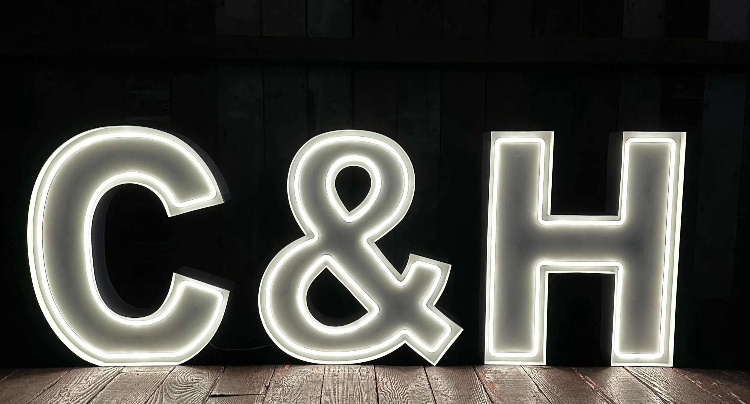 Backlit decorative letters spelling 'C&H' on a dark wall, on a wooden floor.
