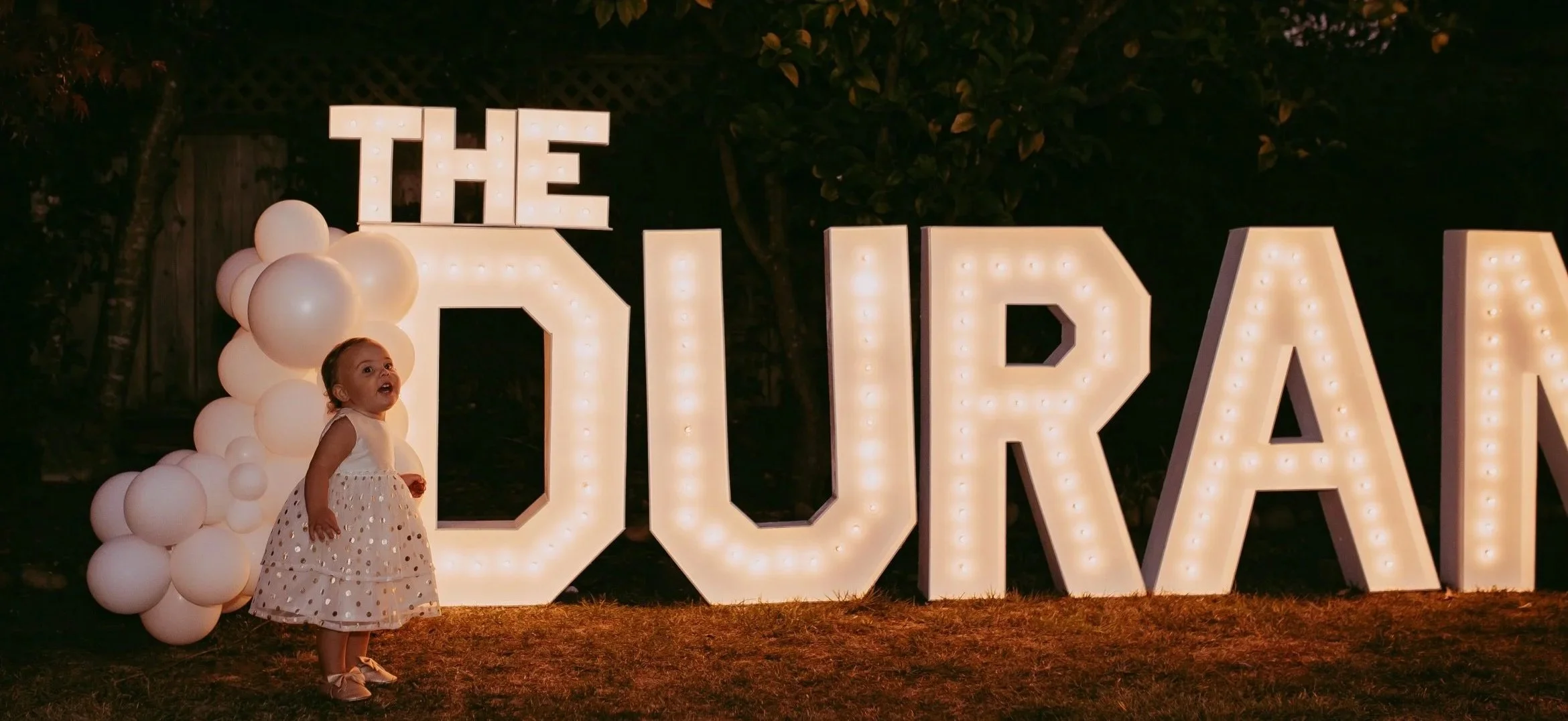 A young girl in a white dress standing in front of large illuminated marquee letters spelling 'TOUR'. The scene is outdoors at night, with balloons assembled on the left side of the girl.