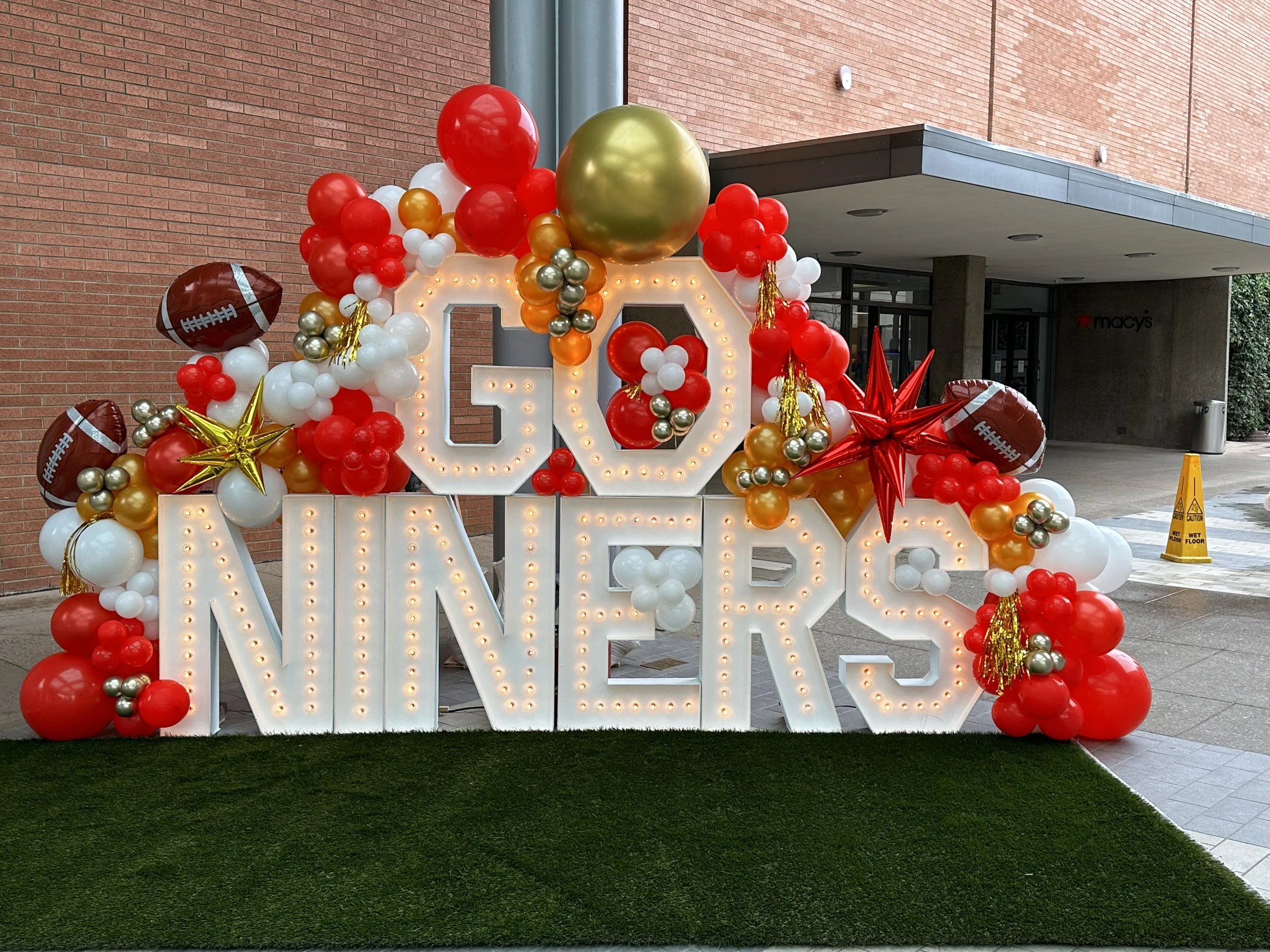 Decorative display with large illuminated text cash reading 'GO NEVER' surrounded by red, white, gold, and metallic balloons, some shaped like footballs, stars, and flowers, set outdoors near a brick building.