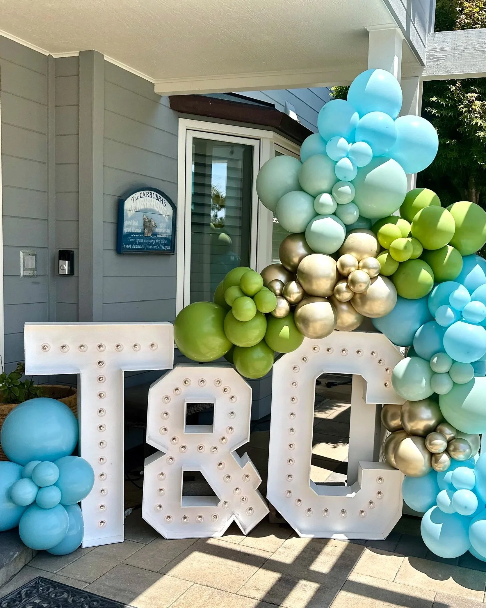 Large white decorative letters 'TRO' with light bulbs, surrounded by blue, green, and gold balloons, set outside a house near a mailbox and window.