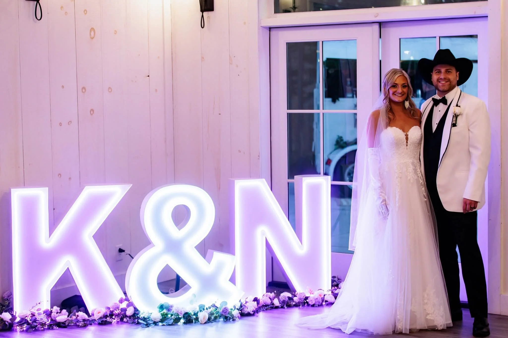 Bride and groom in wedding attire standing beside large illuminated letters 'K&N' with flowers at the base, inside a decorated venue.