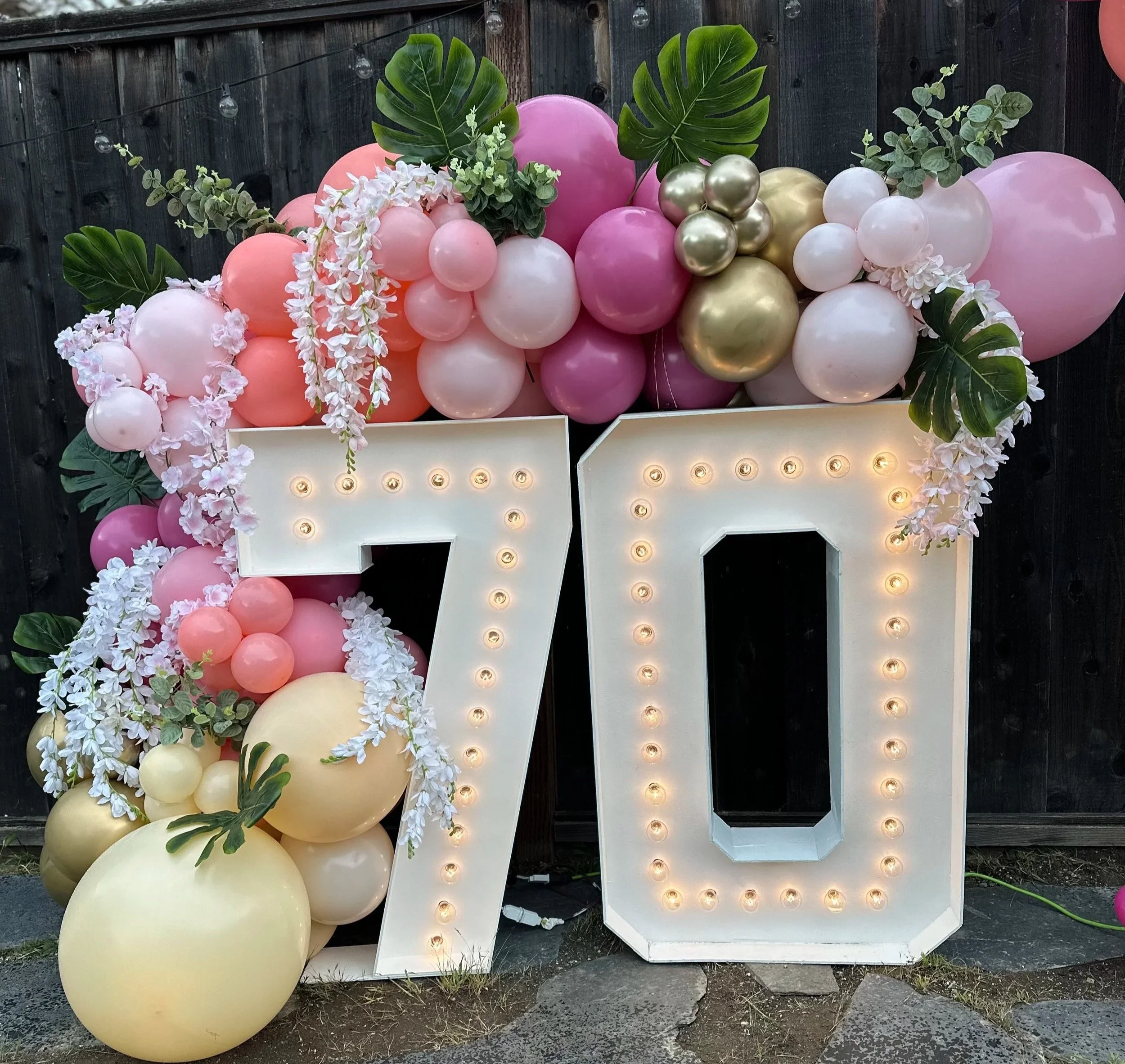 Large illuminated number seven and zero decorated with pink, white, gold balloons, green leaves, and white flowers, likely for a celebration.