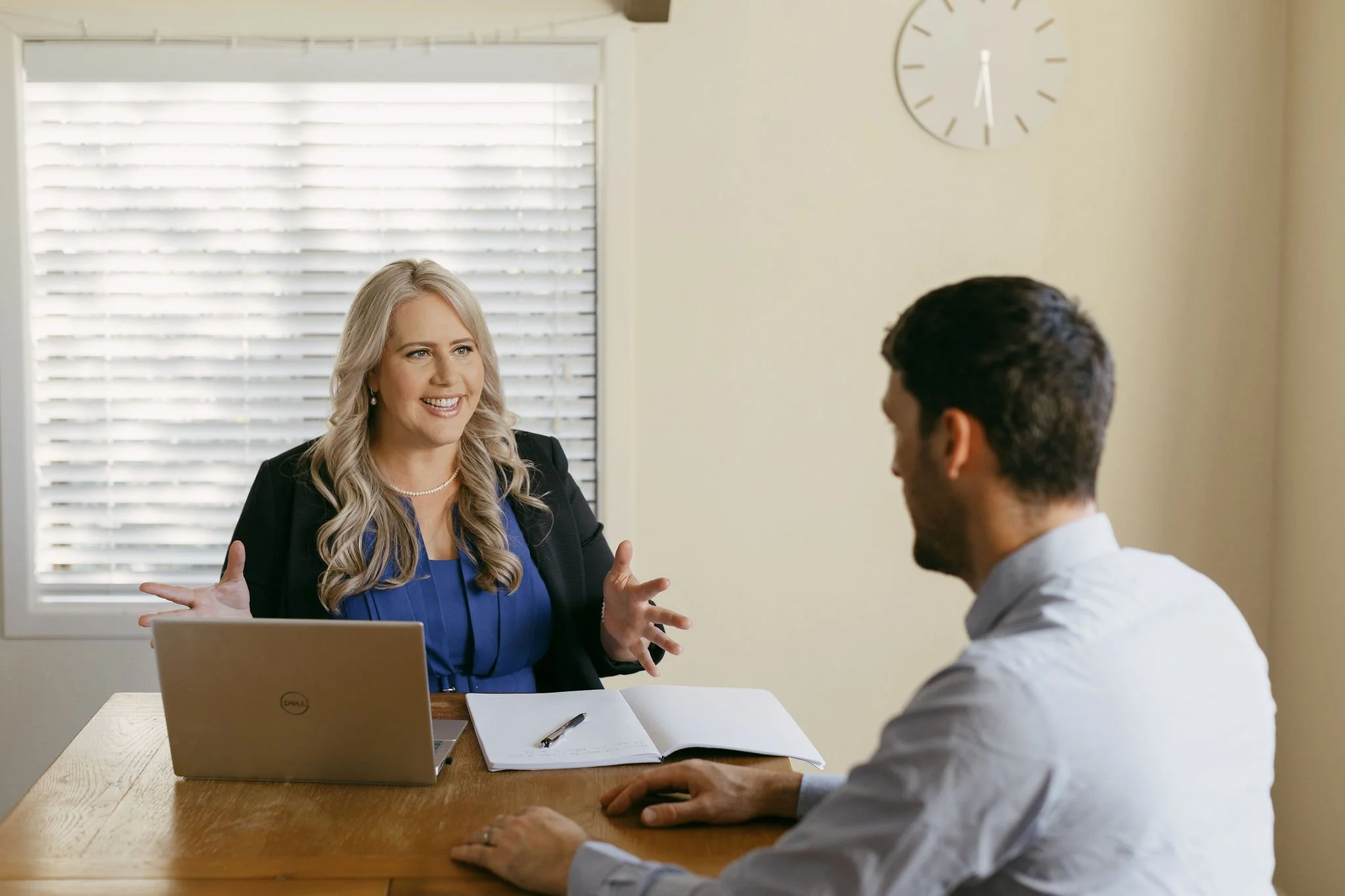 Woman with blonde hair speaking to man with dark hair during a business meeting in an office with beige walls, a large window with blinds, a clock, a laptop, a notebook, and a pen on the table.