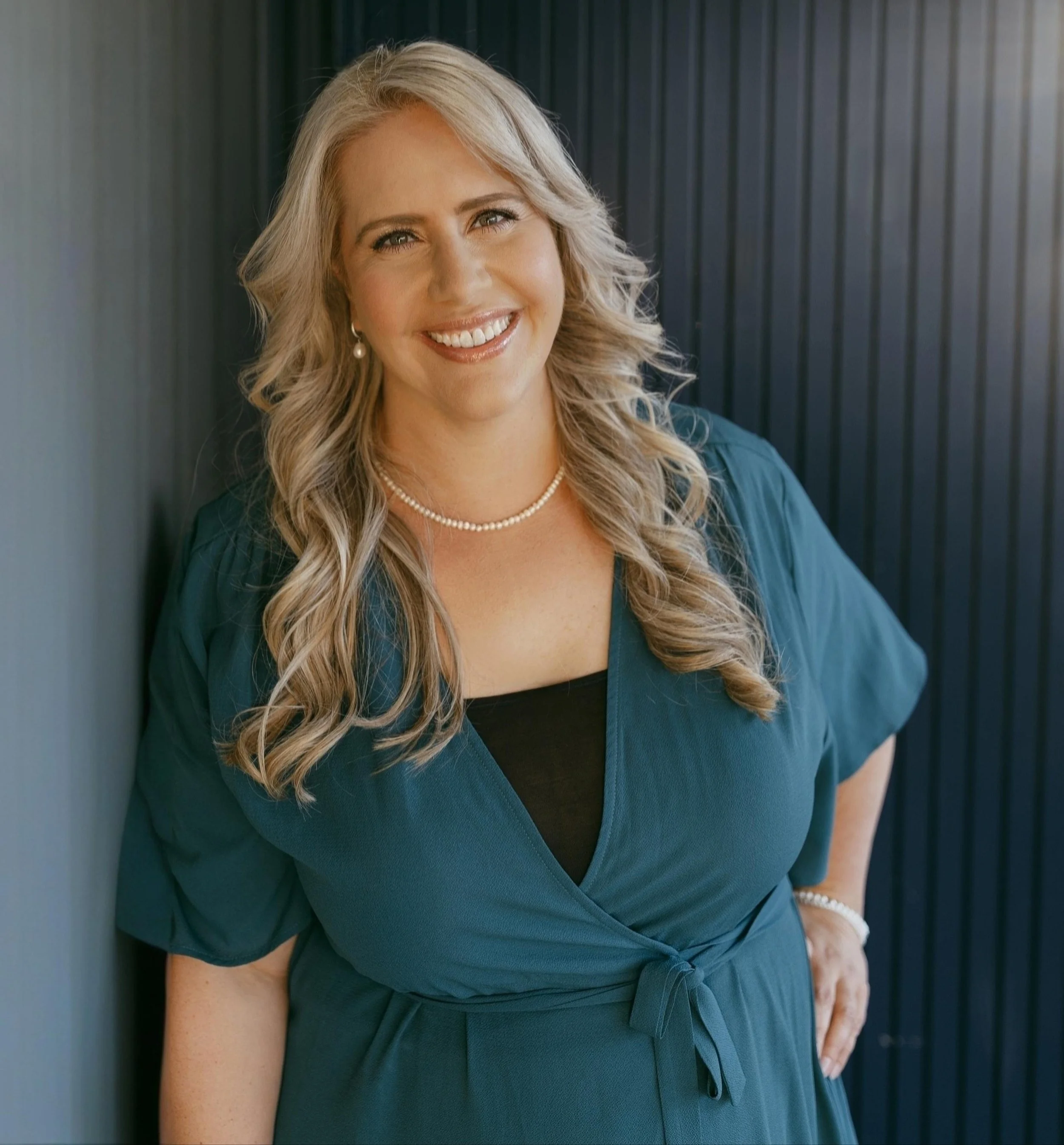 A smiling woman with blonde hair, wearing Pearl earrings, a pearl necklace, and a teal dress, standing against a dark vertical panel wall.