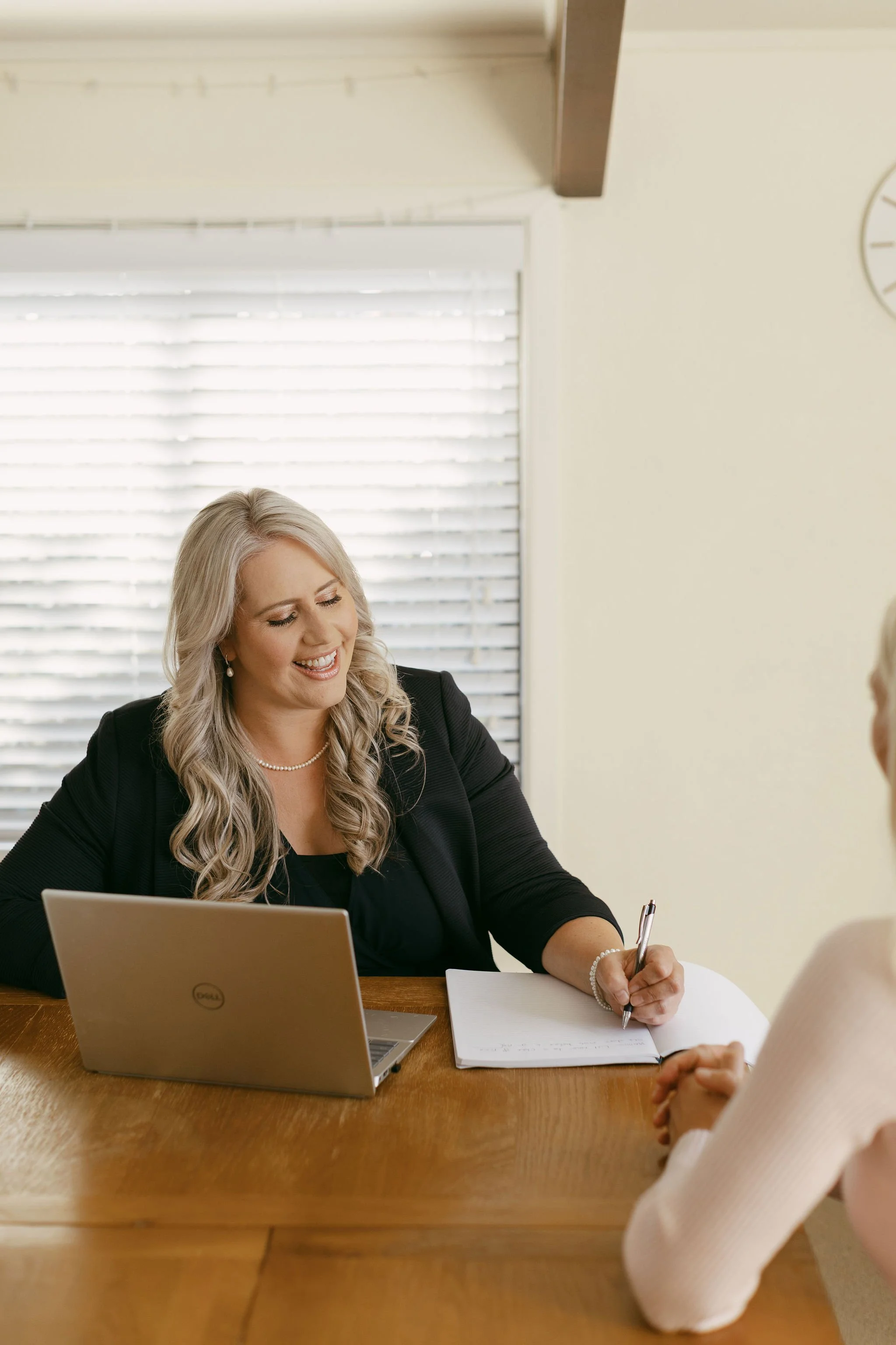 A woman with long blonde hair, wearing a black blazer and a pearl necklace, is smiling and taking notes during a meeting. She has a laptop in front of her on a wooden table, and another person is sitting across from her, partially visible. The room has beige walls, a window with blinds, and a clock on the wall.