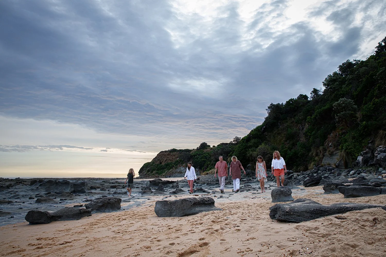 Group of six people walking along a rocky, sandy beach near a hillside with green foliage during cloudy weather.