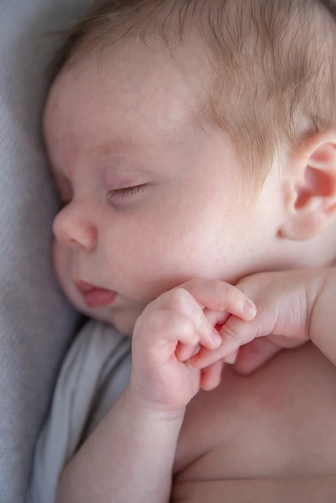 Close-up of a peacefully sleeping infant with light skin, light brown hair, and tiny hands resting near the face.