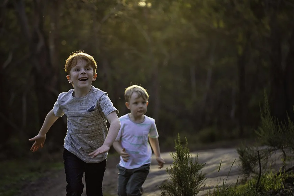 Two young boys running and playing outdoors on a dirt trail through a wooded area, with sunlight filtering through the trees.