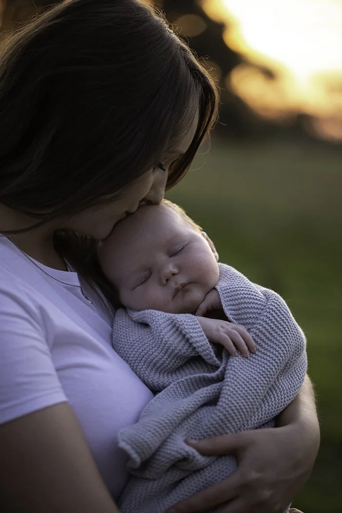 A woman holding a sleeping baby outdoors during sunset, with her head gently resting on the baby's head.