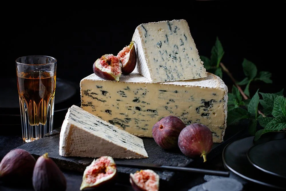 A wedge of blue cheese with a larger block of blue cheese and a smaller piece on a black platter, surrounded by fresh figs and a glass of dark beverage on a dark surface with green leaves in the background.