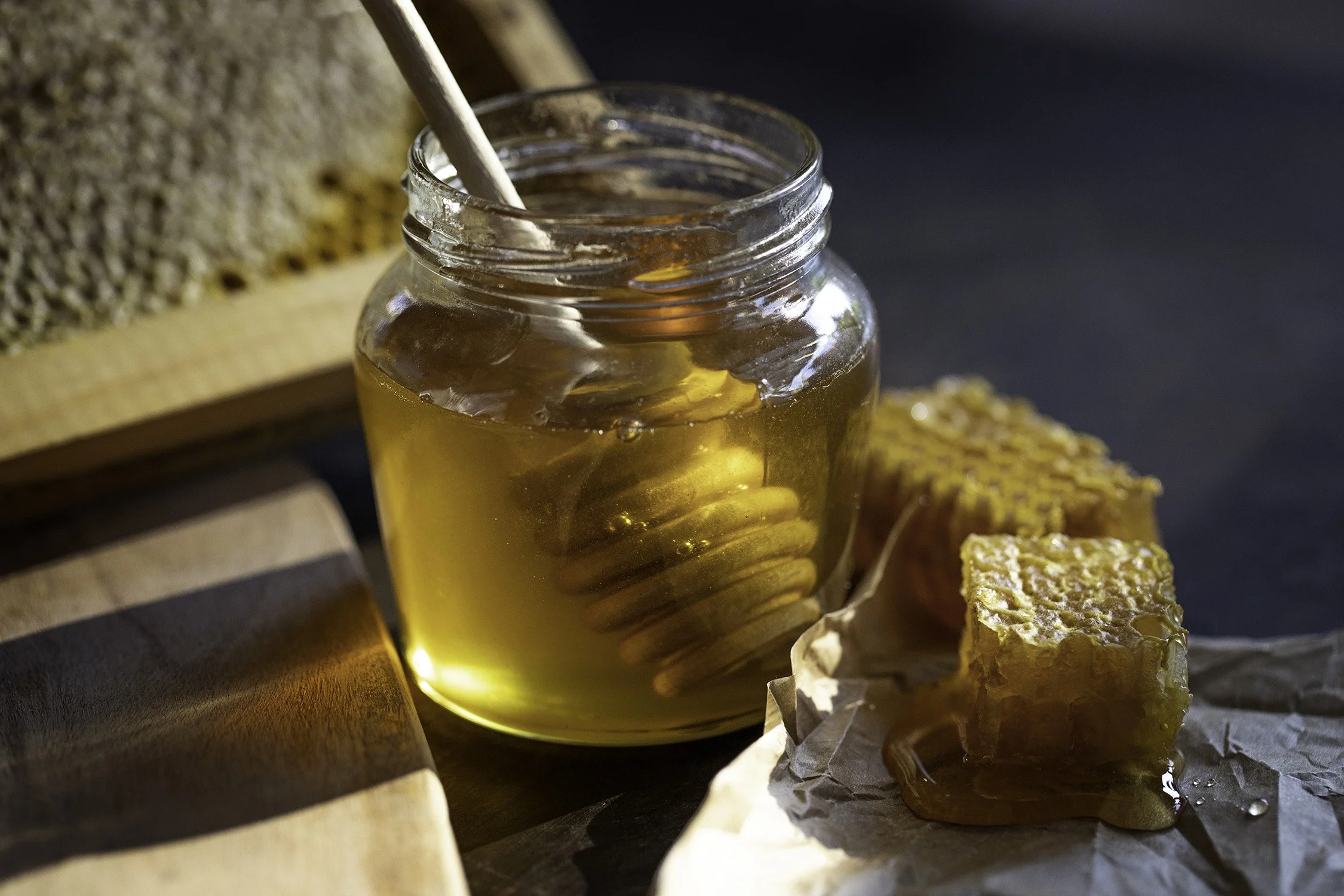 A jar of honey with a honey dipper inside, placed on a wooden surface with honeycomb pieces nearby.