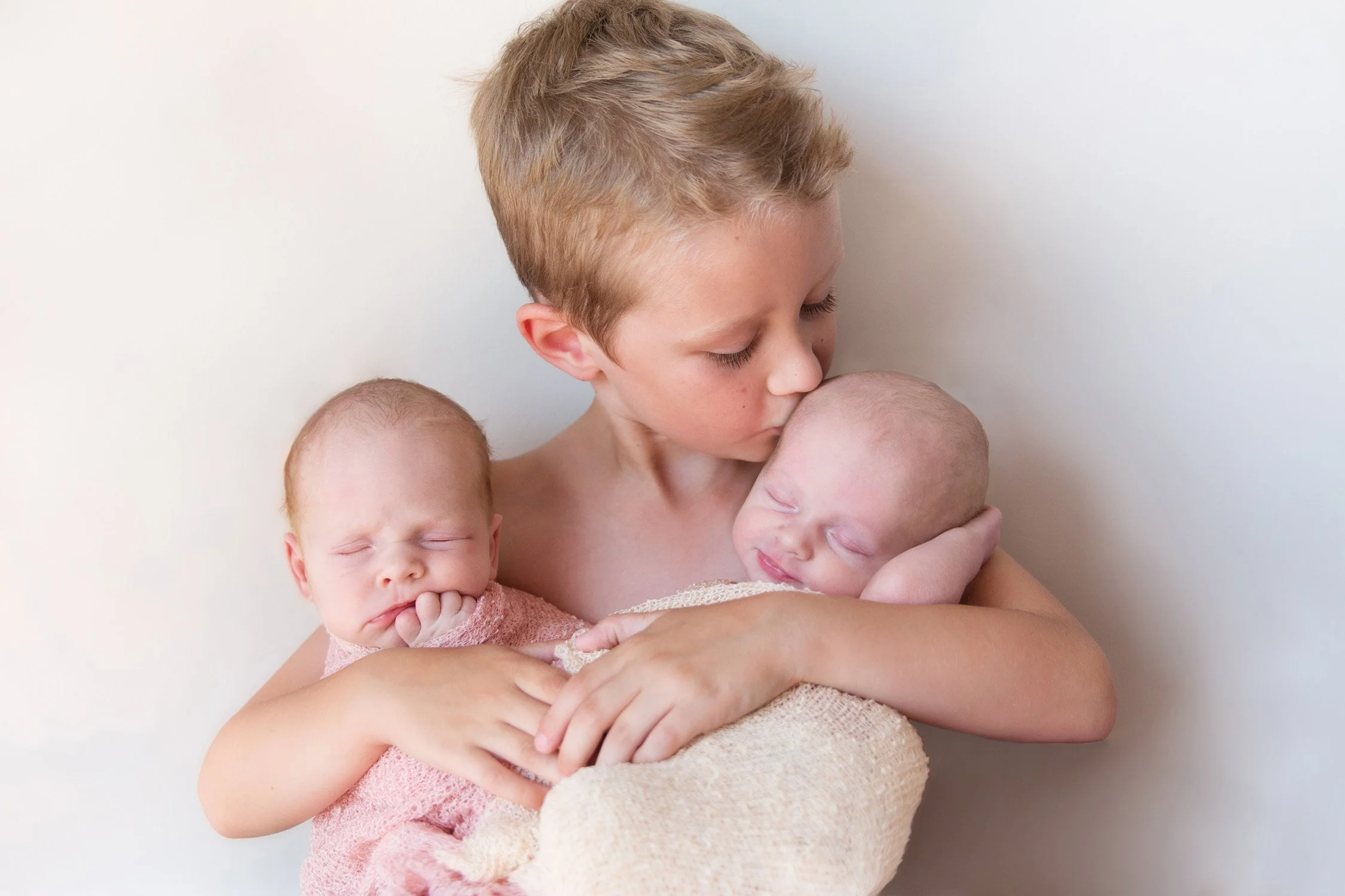 Young boy holding two sleeping babies, one in each arm, gently kissing the baby on the right. The other baby is resting with a hand near the mouth, wearing a pink outfit.