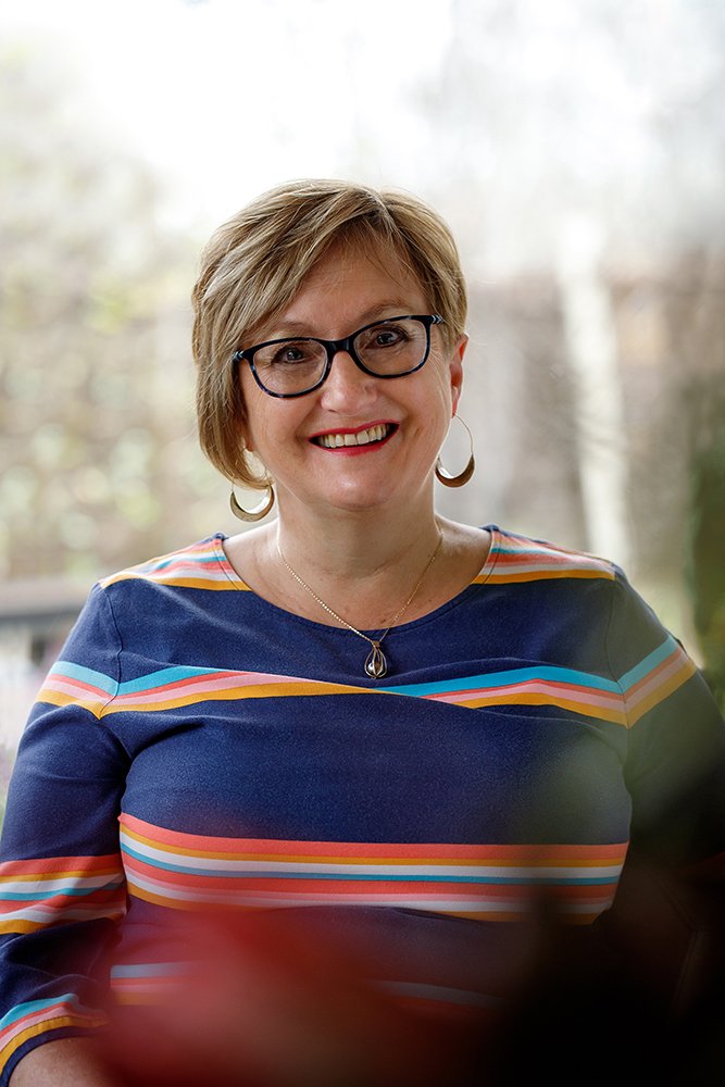 A woman with short blonde hair, glasses, and hoop earrings smiling wearing a colorful striped top with blue, orange, and pink stripes, and a pendant necklace, standing outdoors with blurred trees in the background.