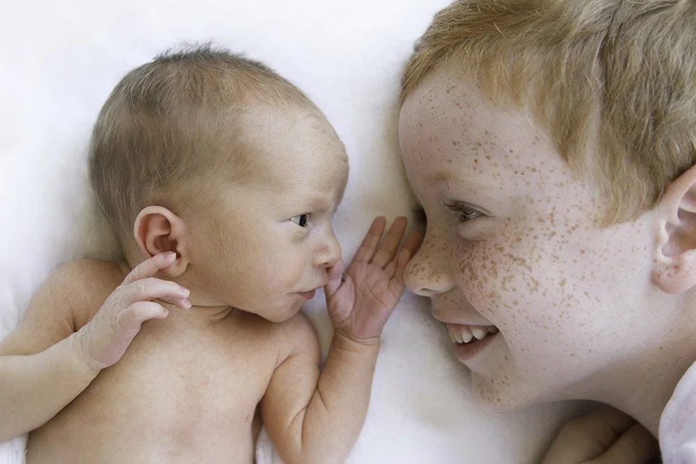 A young boy with freckles and red hair smiling at a small baby with light skin and light hair, as they lie on a white surface, face to face.