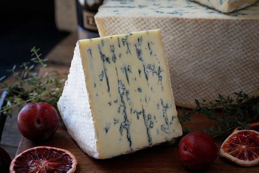 A wedge of blue-veined cheese with a white rind, surrounded by red apples and slices of blood orange on a wooden surface decorated with herbs.