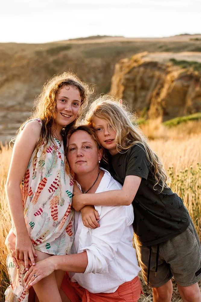 A woman with two young girls in a field during sunset with cliffs in the background.