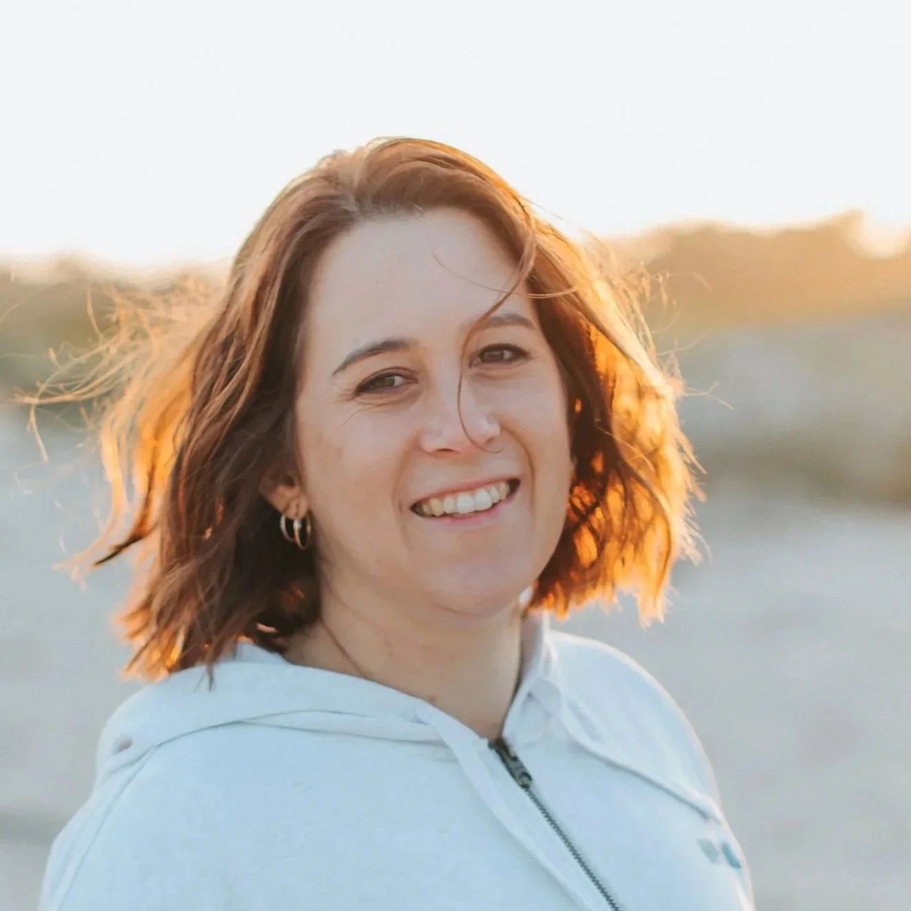 Smiling woman with shoulder-length reddish-brown hair, wearing a white hoodie, standing outdoors at sunset near the beach.
